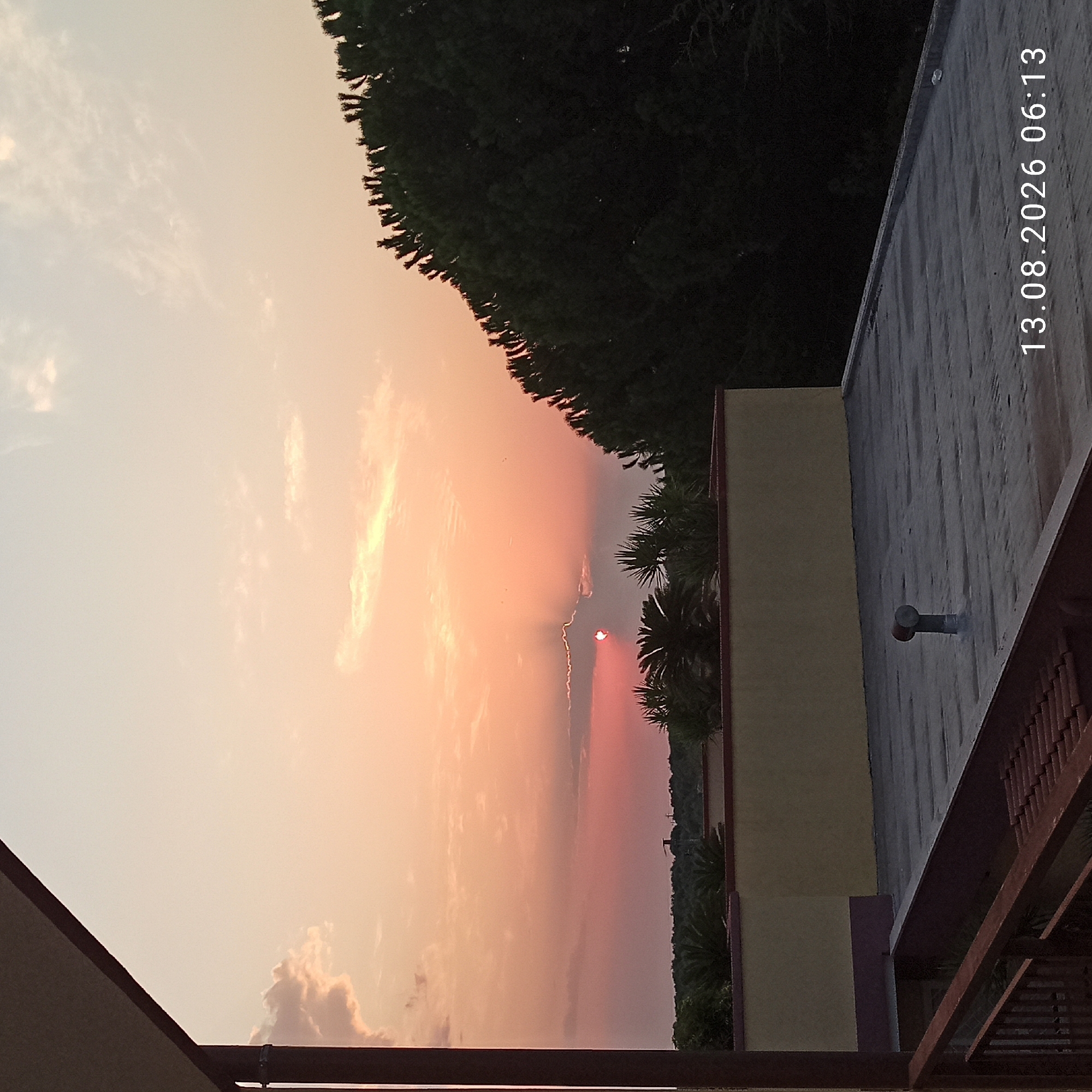 The image depicts a sunset over a rooftop in Bitonto, Italy. The sky is filled with warm hues of orange and pink, with scattered clouds. In the foreground, a flat rooftop is visible, with a vent pipe. The middle ground shows a building with a yellow facade and a red trim, partially obscured by palm trees. Behind the building, a dense line of dark green trees stretches across the horizon. The image is dated 13.08.2026 06:13. No people are visible in the scene.