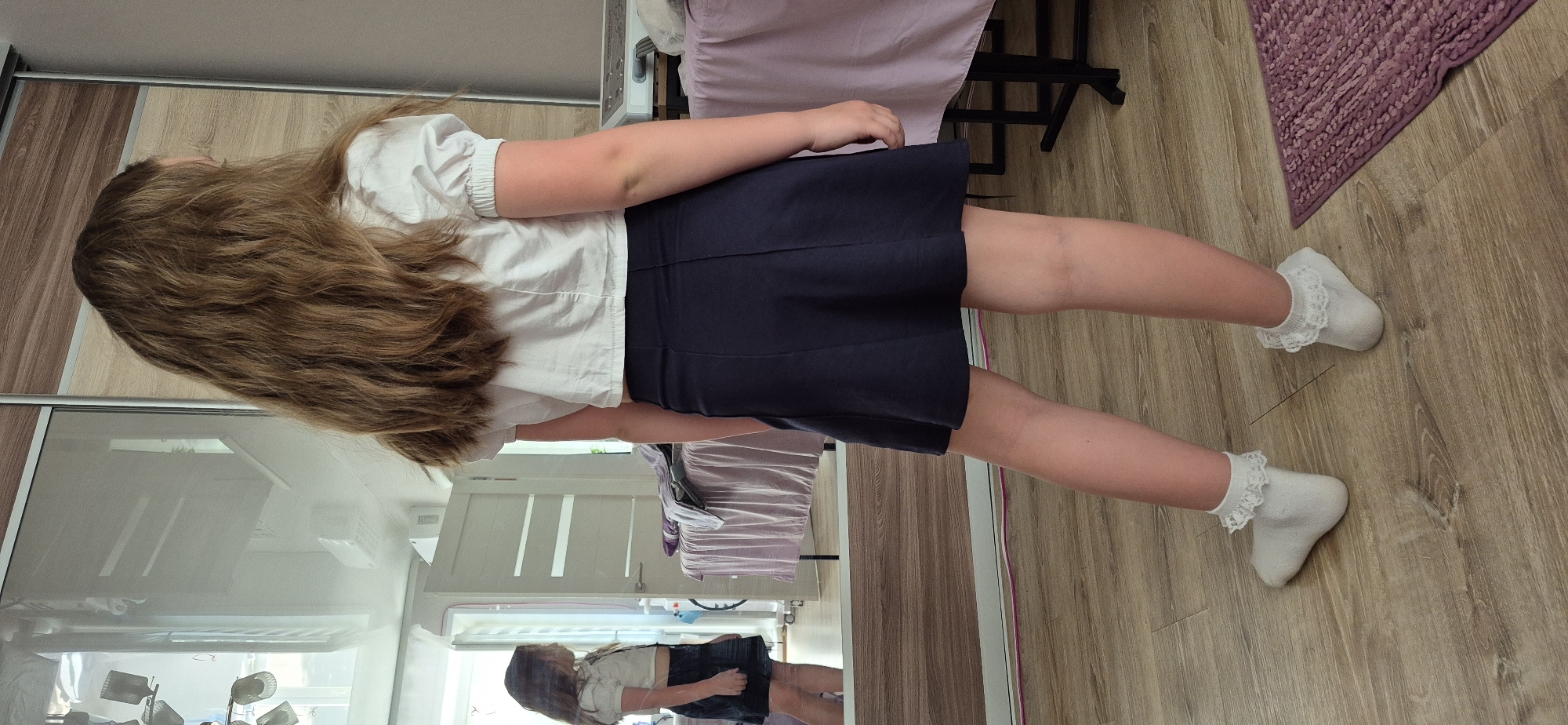 A full-length shot shows a girl wearing a white collared short sleeve shirt and a dark blue skirt standing on a light brown, wood-patterned floor, with her long brown hair cascading down her back. The girl is wearing white ruffled socks. A light purple rug lies to the right, near her feet. In the background, a light purple draped object is on a black-legged surface. Behind the girl, a mirrored wardrobe reflects the room and a smaller image of the girl herself. The setting appears to be inside a home, though the exact location within the city of Unknown, Unknown is unspecified.