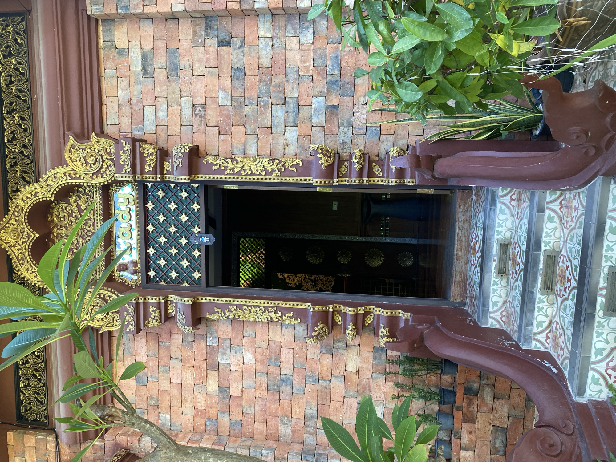 This is a close-up, eye-level shot of the entrance to a building in Baan Lamai, Thailand, likely a temple or shrine, given the ornate architectural style. The entrance features a deep red wooden door frame with intricate gold embellishments and a decorative archway above. The archway has a golden, scroll-like design and a panel with a green lattice pattern overlaid with small, golden diamond shapes. Above the lattice, there are colorful, stylized letters in a script that appears to be Thai.
The building's exterior is constructed of rustic, reddish-orange bricks, with some variations in color and texture. To the left of the entrance, a bare-branched tree with lush green leaves is partially visible. To the right and in front of the entrance, potted plants and greenery add to the tropical atmosphere.
A set of steps leads up to the open doorway. The steps are covered with patterned tiles, featuring a repeating floral design in green, red, and yellow. The sides of the staircase are adorned with sculpted, dark red, curving banisters that have a mythical, perhaps dragon-like, appearance.
Inside the doorway, the interior is dimly lit, revealing dark wooden panels with intricate gold designs and what appears to be a dark vase or object in the far background. The overall impression is one of detailed craftsmanship and traditional Southeast Asian aesthetics.