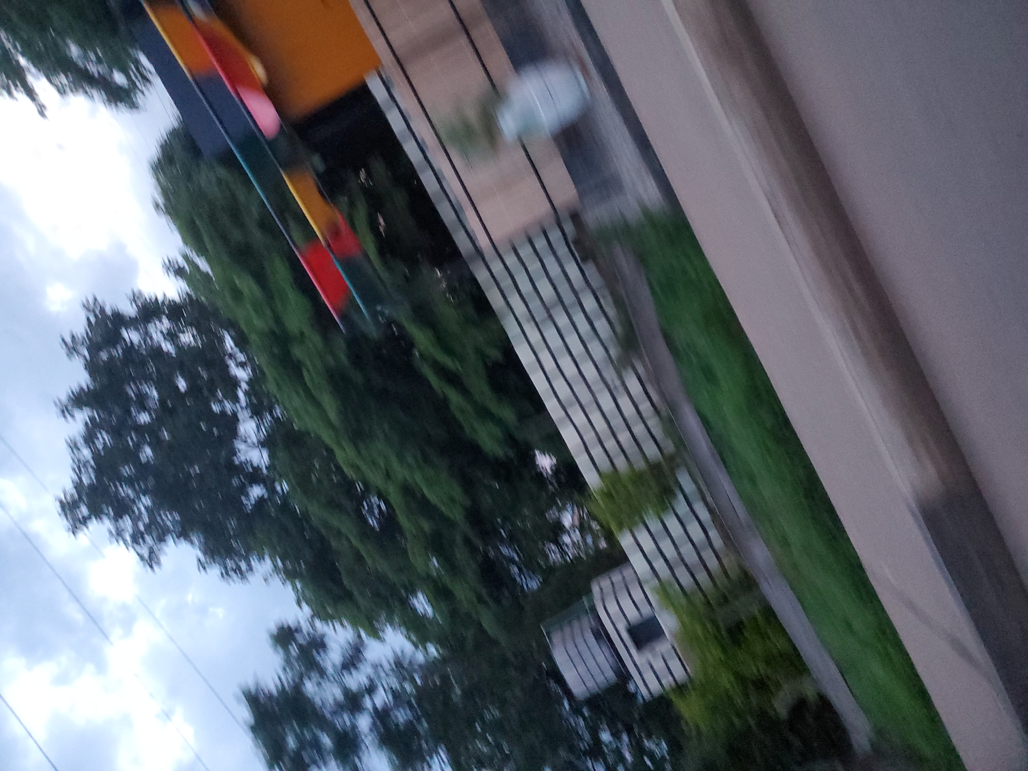 The image depicts a street scene in Gura Top, Nigeria, captured with motion blur. A section of a yellow structure with colorful horizontal bands of red, yellow, green, and blue is visible at the top right. Below this, a white fence with black horizontal lines stretches across the frame. To the left of the fence, there is a building with a water tank on top, and several small trees. The ground in front of the fence is covered with grass. In the foreground, a portion of a road is visible. The sky is cloudy.