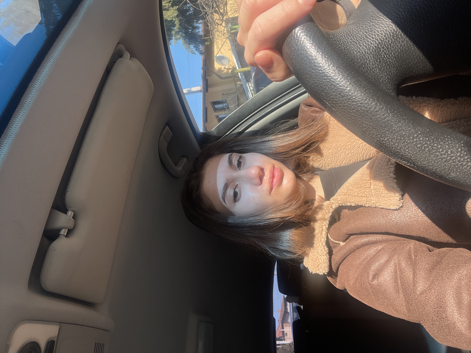 A young adult female is in the driver's seat of a car. She is holding the steering wheel with her left hand. She is wearing a brown jacket with a fur collar. The sun visor on the driver's side is down. Through the windshield, buildings are visible. Based on the architecture and vegetation visible through the windshield, the location is likely Sofia, Bulgaria.