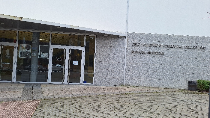 The image displays the exterior of the Centro Cívico e Cultural de Arteixo Manuel Miguélez in Arteixo, Spain. The building facade features a light-grey, textured wall with raised silver lettering identifying "CENTRO CÍVICO E CULTURAL DE ARTEIXO" and "MANUEL MIGUÉLEZ." The entrance consists of multiple glass double doors with white frames, recessed under a white overhang. Various white and dark notices and posters are affixed to the glass panels and an interior structural column. Reflections of a light-colored vehicle and interior elements are visible in the glass. The ground in front of the building is paved with interlocking concrete blocks in grey and reddish-brown tones, with a darker concrete path directly preceding the entrance. To the far right, a dark-colored cylindrical trash receptacle is situated next to a small, trimmed green shrub. No individuals are present in the scene.