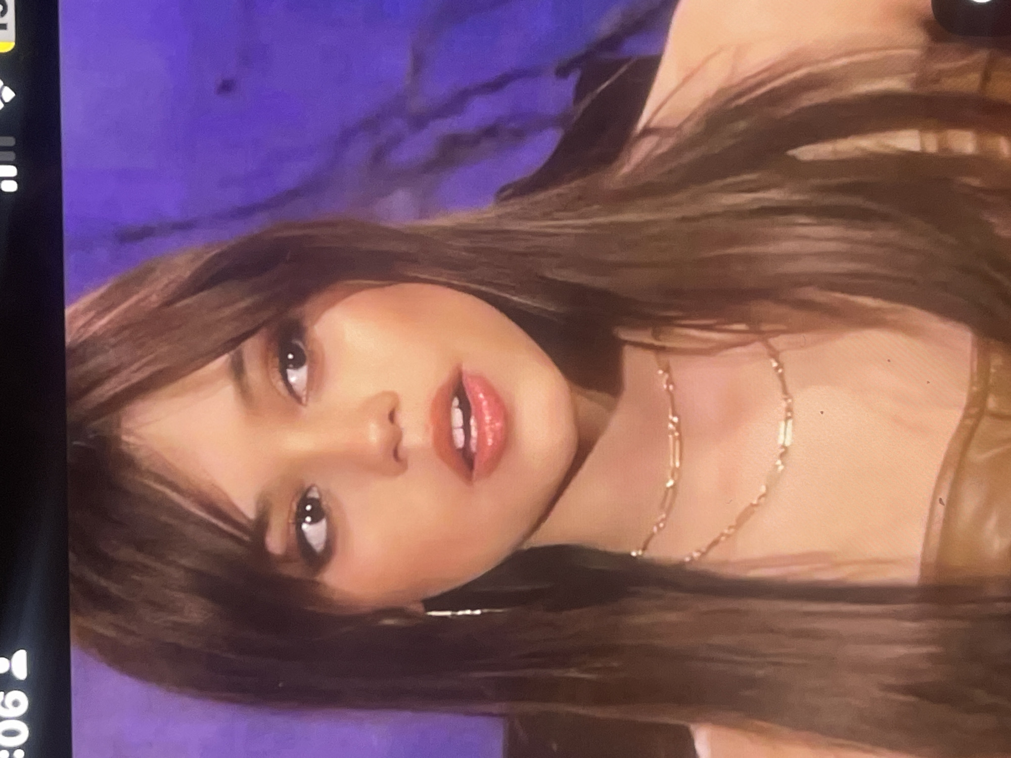 A close-up portrait of a young woman, rotated 90 degrees clockwise, captures her looking directly at the viewer. She has long, dark brown hair with bangs that fall across her forehead, and her lips are slightly parted, revealing some teeth, enhanced by what appears to be pink lipstick. Her expression is neutral to slightly alluring. She is wearing a delicate gold-toned necklace with small, rectangular links. A light brown or tan garment is visible on her shoulder and chest. The background is a solid, textured purple, suggesting an indoor setting like a studio or a room with a distinct wall color. The image itself appears to be a photograph taken of a digital screen, indicated by a black bar on the left side that displays mobile device interface elements. These elements include icons for full signal strength, Wi-Fi connectivity, and a full battery. The time "9:06" is also partially visible on this bar. No specific location cues for Poltava, Ukraine, are discernible within the image.