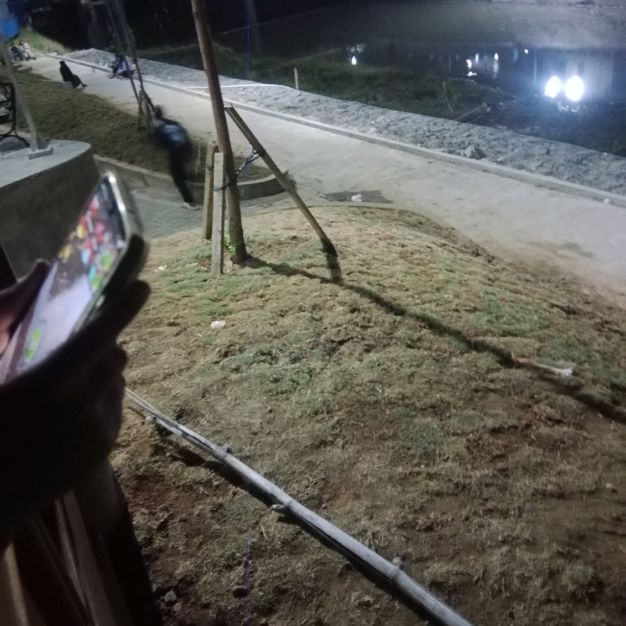 The image depicts a night scene near a body of water, possibly a lake or river, in Tlogorejo, Indonesia. In the foreground, a person's hand is holding a smartphone, its screen displaying colorful app icons. The phone is silver and appears to be a modern model. To the left, a concrete structure with rounded edges is visible. The ground is covered in dry grass and dirt, with a metal pipe running diagonally across it. In the mid-ground, a paved walkway runs along the edge of the body of water, which reflects distant lights. Several people are present on or near the walkway. One individual, wearing dark clothing, is walking away from the camera. Another person, in dark clothing with a blue element, appears to be leaning or sitting. Further back, two individuals are seated on the walkway. The environment suggests a public space, possibly a park or promenade. The lighting indicates it is nighttime, with artificial lights contributing to the illumination and reflections in the water.