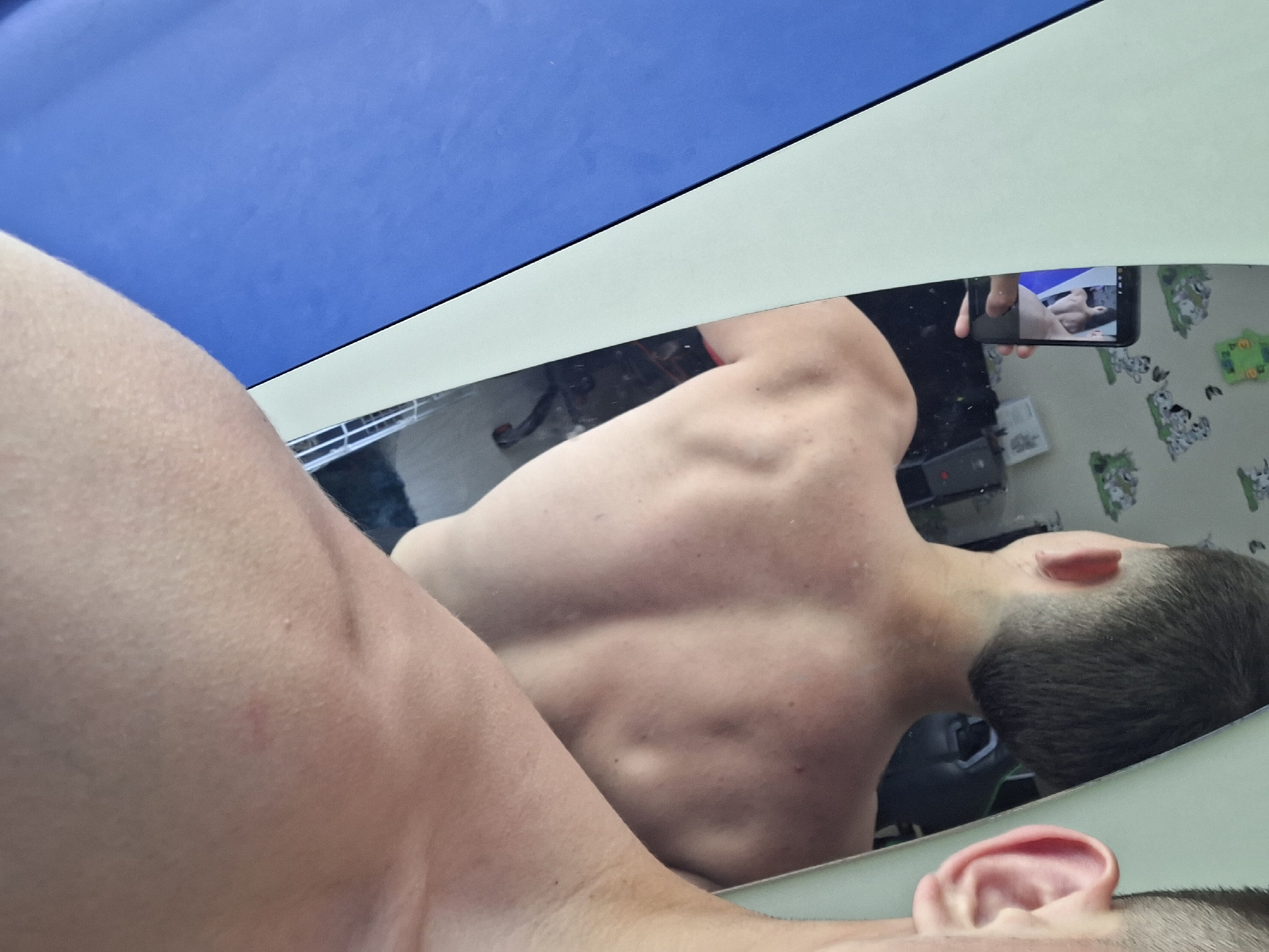 This is a photograph of a young person's back and shoulders, taken in the city of Varna, Bulgaria. The subject is shirtless, and their musculature is visible. The image is taken in a room, as evidenced by the wallpaper with cartoonish patterns in the background and a mirror reflecting the scene. A smartphone is visible in the mirror, held by the subject, indicating a selfie is being taken. The room appears to be a bedroom or a personal space, with a blue curtain or fabric visible on the left side of the frame. The lighting is natural, suggesting daylight.