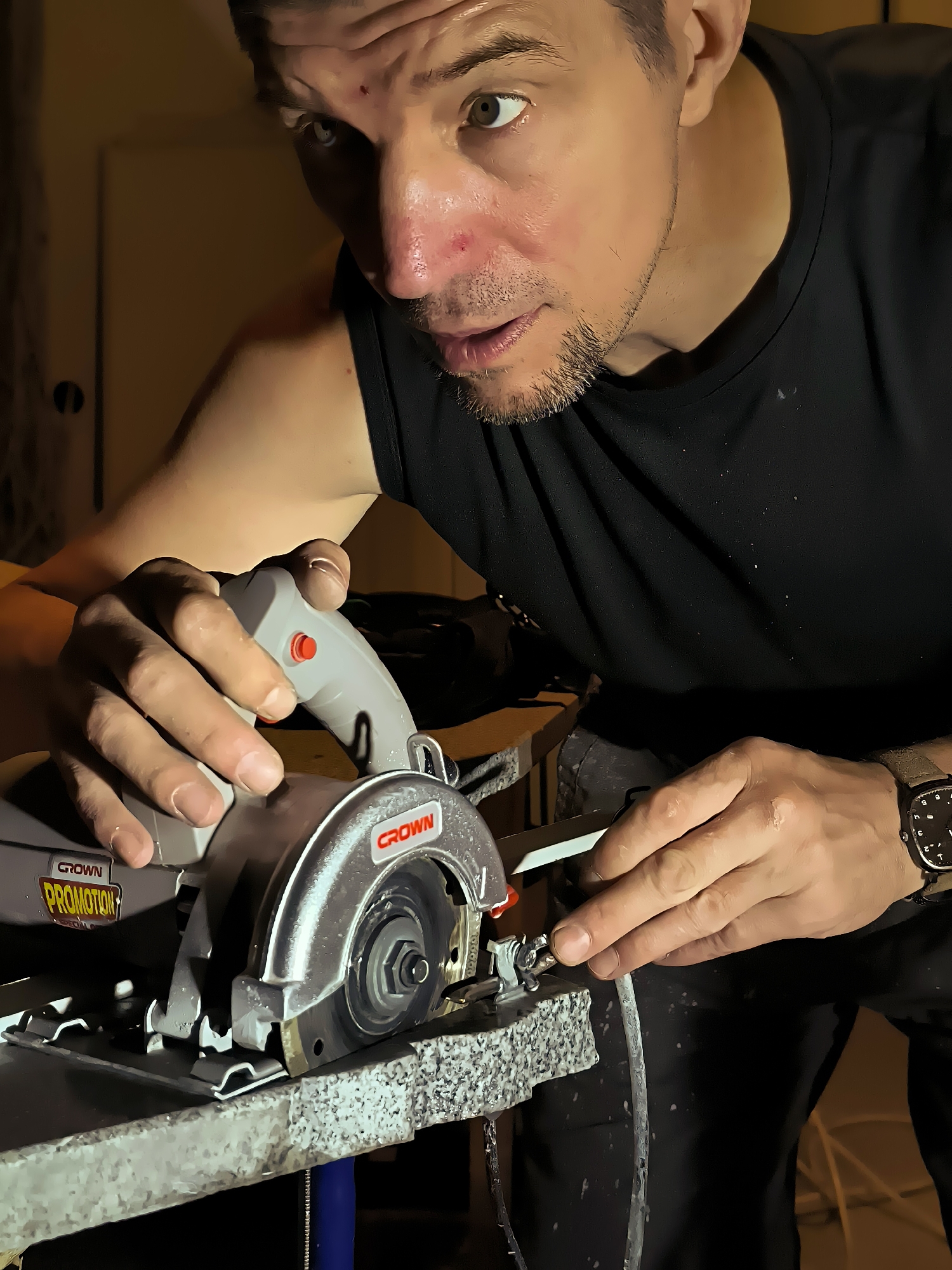 A man is using a circular saw to cut a piece of material. He is wearing a black tank top and dark pants. The circular saw is gray and red, with the brand name "CROWN" visible. The man's face is visible in the upper portion of the image; he has short, dark hair and stubble. His eyes are open and he appears focused on his task. He is wearing a watch on his left wrist. The background is dimly lit and appears to be an indoor workspace. There is no indication of the location in the city of Qarağandı city administration, Kazakhstan in the image.