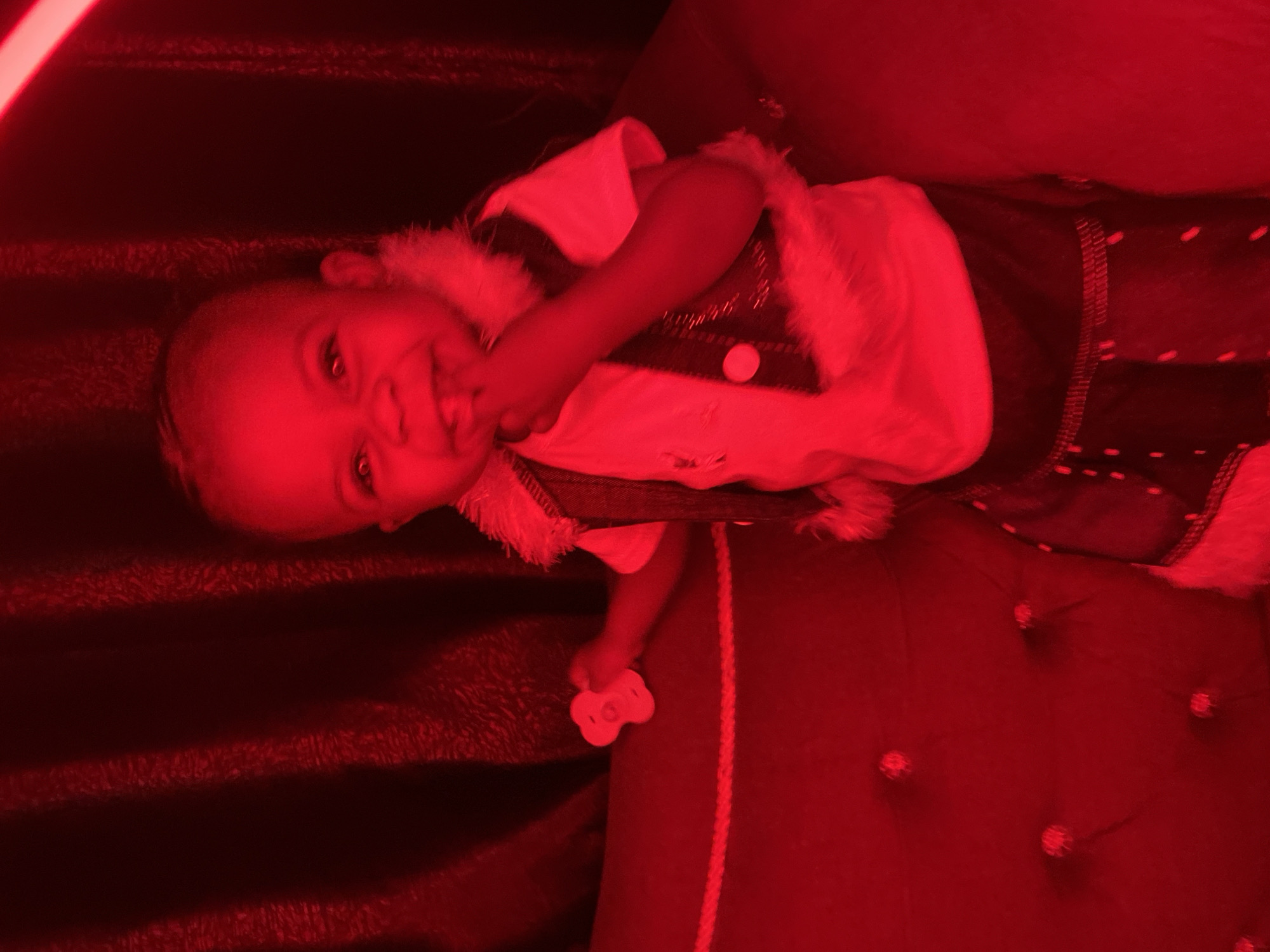 A toddler is seated on a red tufted couch with a dark red, textured backdrop. The toddler is wearing a white t-shirt, a dark vest adorned with fur trim and decorative buttons, and dark shorts with fur trim. The child is holding a small, white, three-petal-shaped object in their left hand and has their right index finger in their mouth. The scene is illuminated by a strong red light, casting a red hue over everything. The location is identified as a residential setting in Abakaliki, Nigeria.