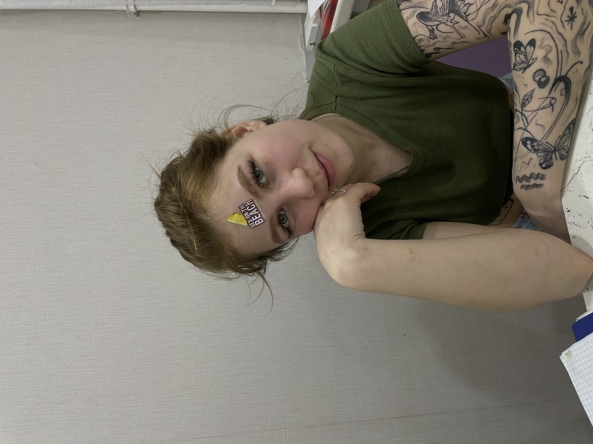A medium shot of a fair-skinned person with light brown hair posing with their right cheek resting on their hand. They wear a green ribbed shirt and a silver necklace, and there is a small sticker on their forehead. The person has multiple tattoos on their left arm. The background is a plain, light gray wall. The location is Марковский, Russia.