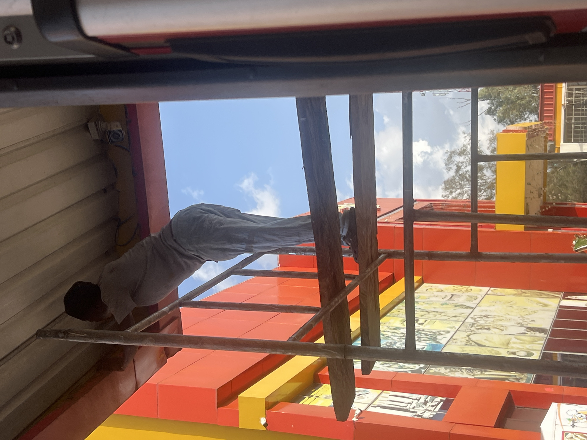 A man in grey clothing is precariously positioned on a metal ladder, reaching towards the roof of a building. The building's exterior is decorated with vibrant red and yellow blocks. A security camera is mounted on the roof near the man. Behind the building, a blue sky with scattered clouds is visible. The scene is likely a work in progress, possibly maintenance or construction. The location is Gura Top, Nigeria.