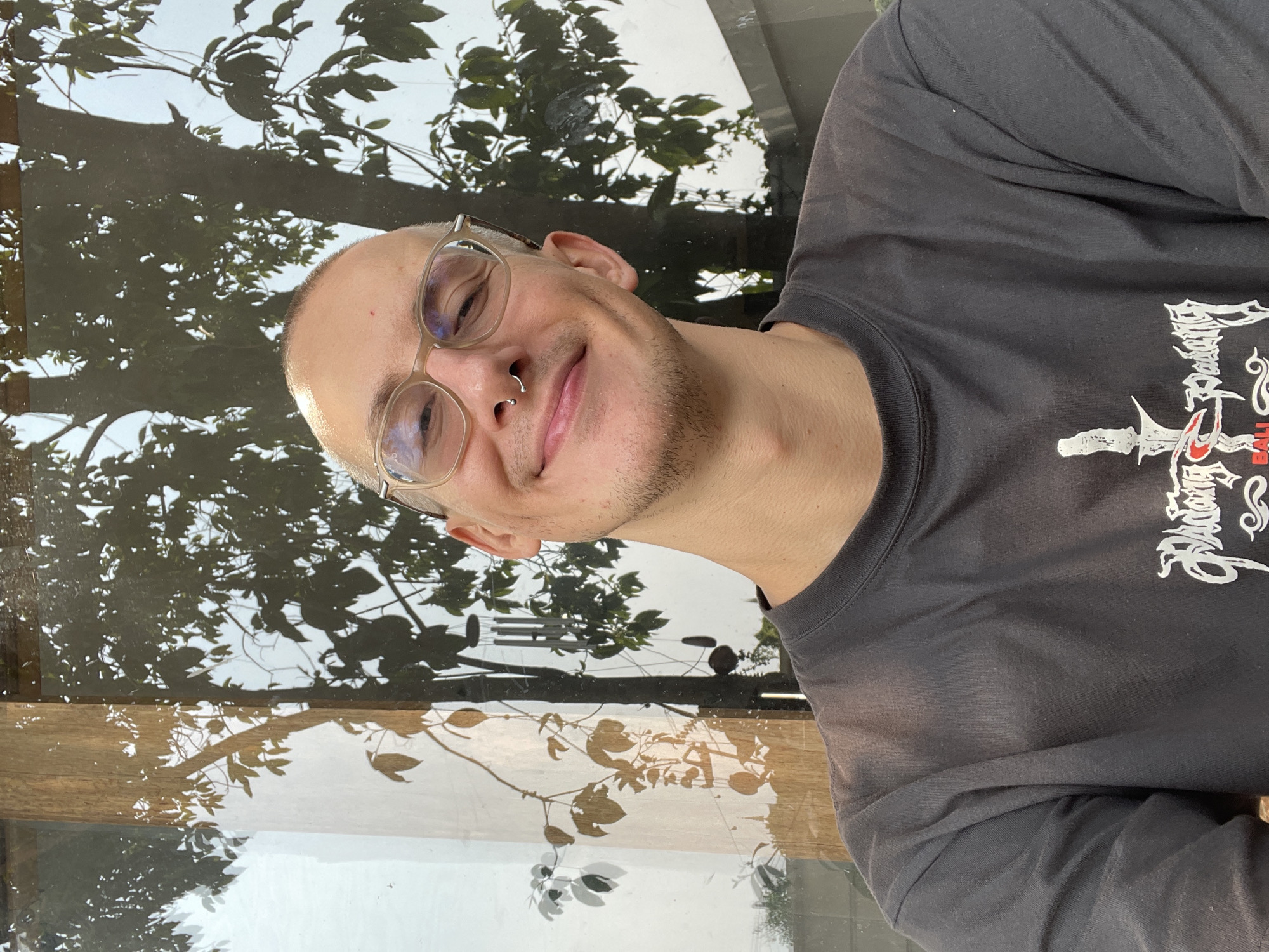 A man with short, light hair, wearing glasses and a nose ring, smiles. He is wearing a gray long-sleeved shirt with a design on the front. The background shows a window reflecting trees and foliage. A wooden frame borders the window. The location is Chang Phueak, Thailand.
