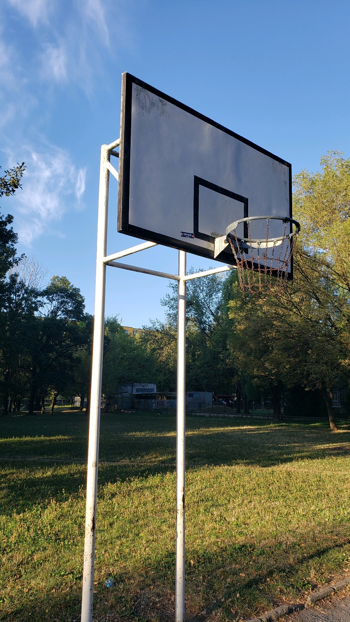 The image shows a basketball hoop and backboard in an outdoor setting. The basketball hoop appears to be made of metal with a chain net. The backboard is rectangular and white with a black border. The hoop is mounted on a white metal frame with two vertical posts. The surrounding environment is a grassy park with trees in the background. The sky is blue with some wispy clouds. In the distance, there are some buildings visible. This scene is located in Prešov, Slovakia, likely at a public park or sports facility. There are no people visible in the image. The ground is covered in grass and some fallen leaves. The lighting suggests it is daytime, likely late afternoon due to the angle of the shadows.