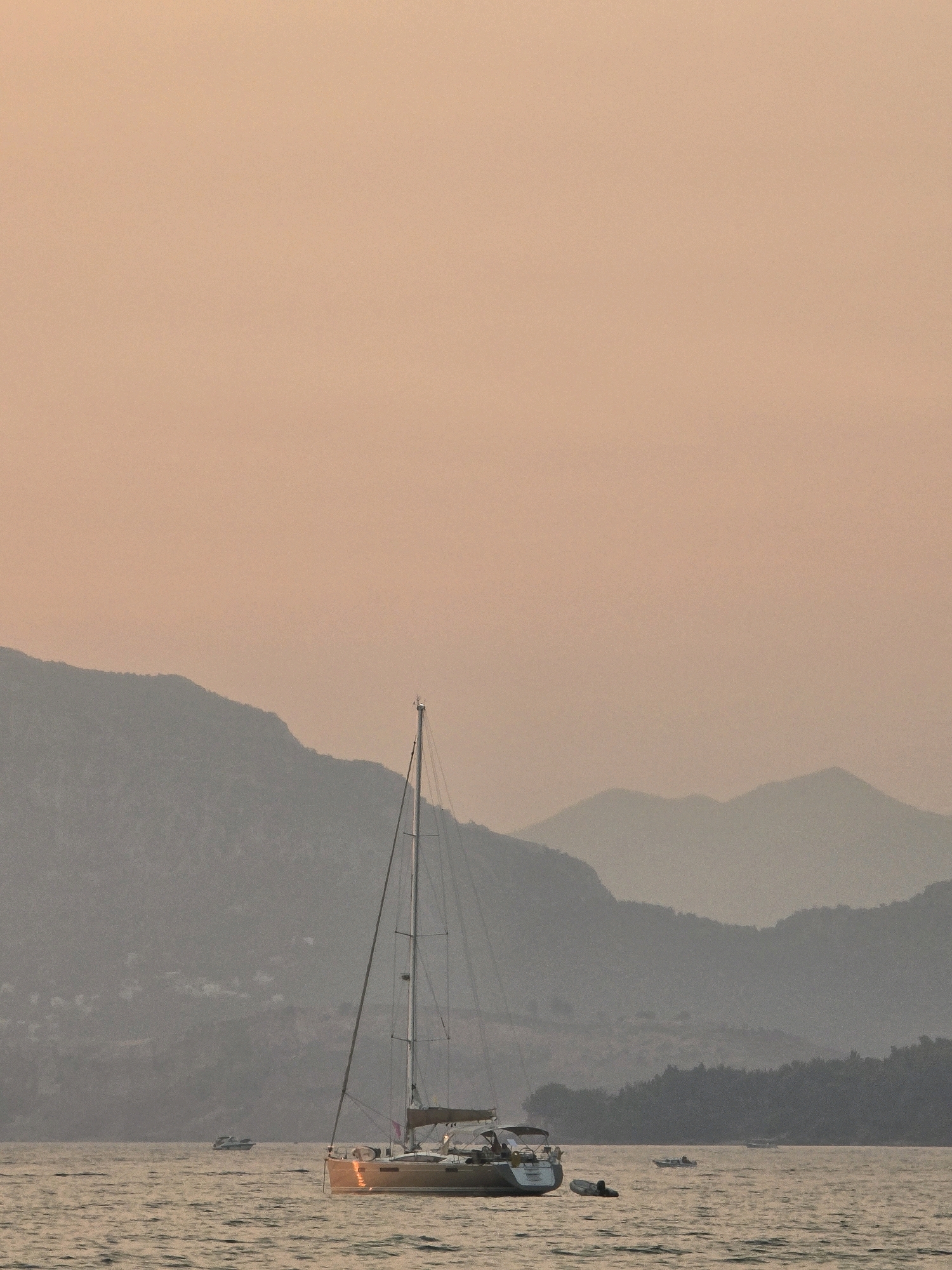 A sailboat is anchored in calm waters, with a hazy, orange sky overhead. The sailboat is the dominant object in the foreground, with its mast extending high into the frame. Several smaller boats are visible in the distance. Rolling hills and mountains form a backdrop, their outlines softened by the atmospheric haze. The scene suggests a serene, late afternoon or early evening on the coast. The image does not contain any recognizable landmarks or specific features that would definitively place it in Bar, Montenegro.