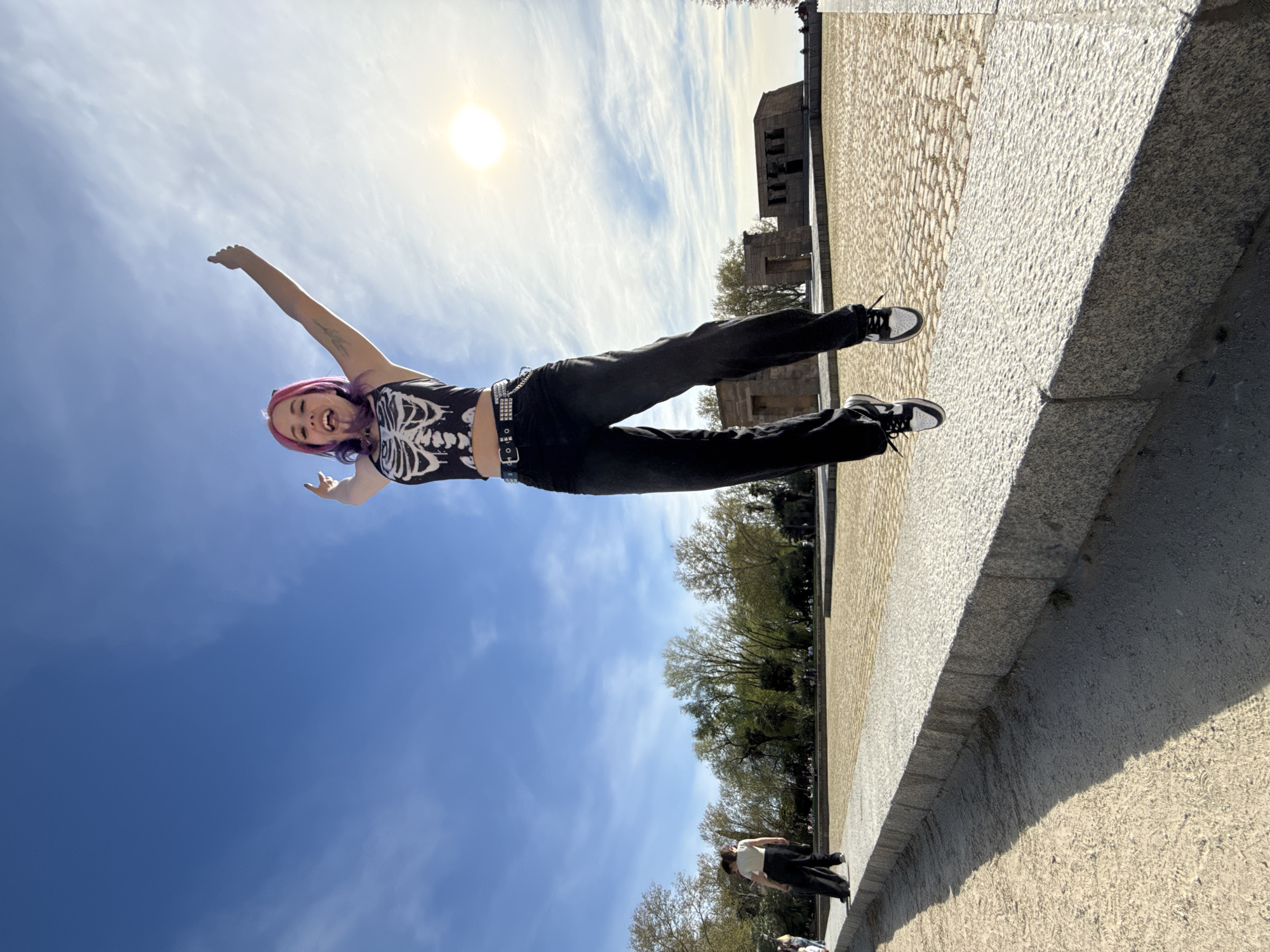 A woman with pink hair is lying on a wall with her arm raised in front of a blue sky. She is wearing a black skeleton t-shirt and black pants. In the background is a building made of stone and trees. There is also a crowd of people walking in the background. The photograph appears to be taken in Madrid, Spain.
