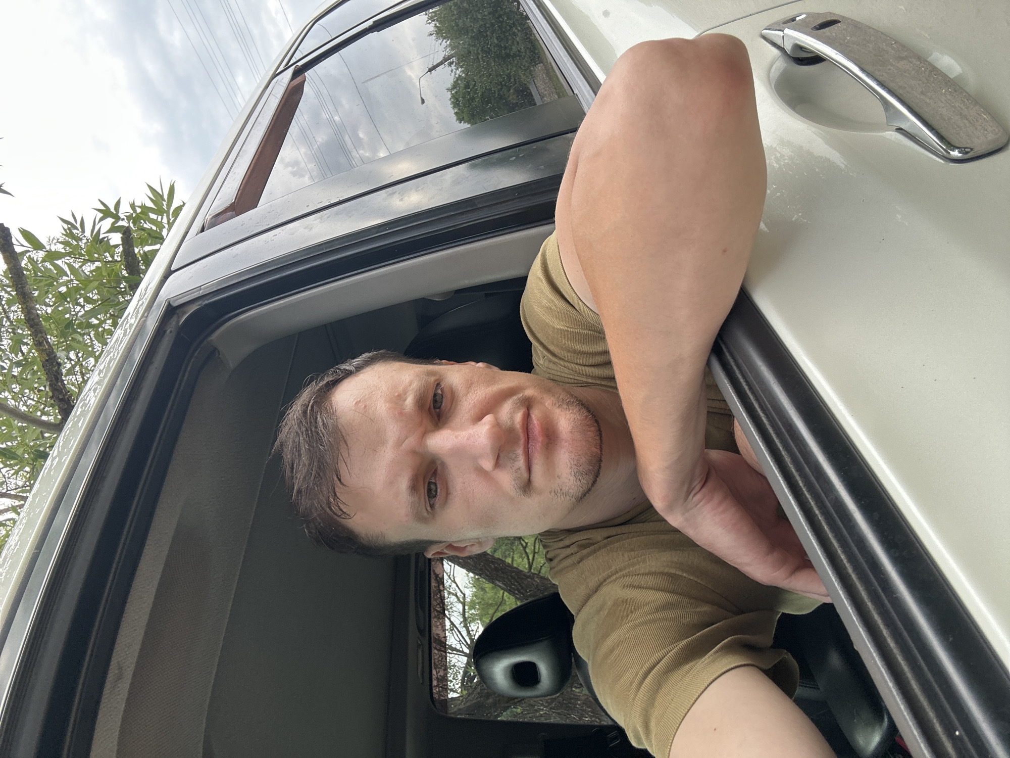 A young Caucasian male is pictured from the chest up, leaning his arm out of a car window. He has short, dark brown hair and is wearing a short-sleeved, olive-green t-shirt. His skin is fair, and he has a neutral expression on his face. The car appears to be white and is parked outdoors, with lush green trees and a cloudy sky visible in the background. The image does not provide enough information to determine the specific location within the city of Makiv, Ukraine.
