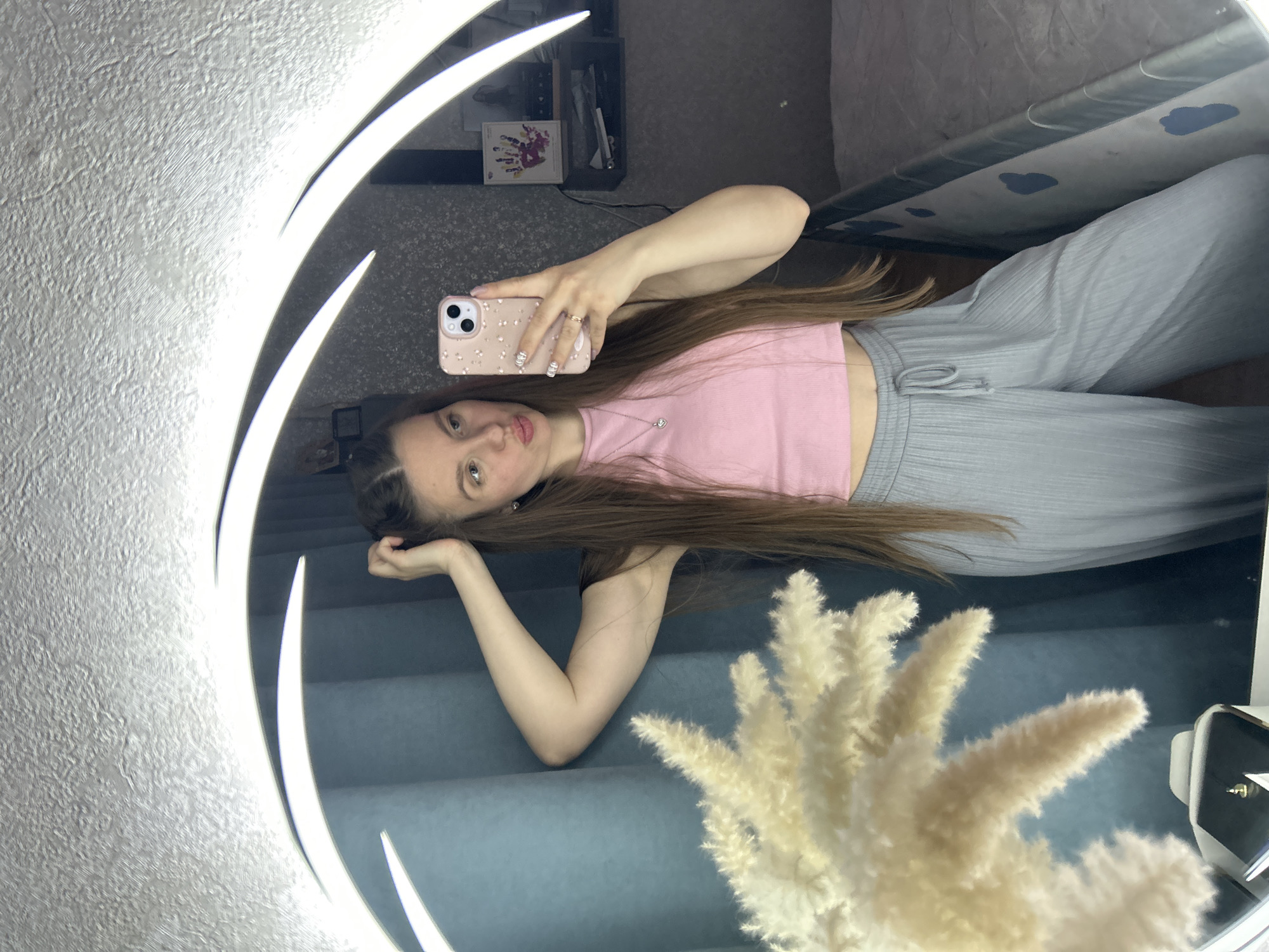 A young woman is taking a selfie in a mirror. She is wearing a pink top and gray pants. Her hair is long and brown. She is holding a phone with a light pink case with flowers. The background of the photo is a bedroom, including a shelf, a bed, and a decorative white plant. The mirror is circular and has a light around it. There is no information in the image to suggest the photo was taken in Klin, Russia.