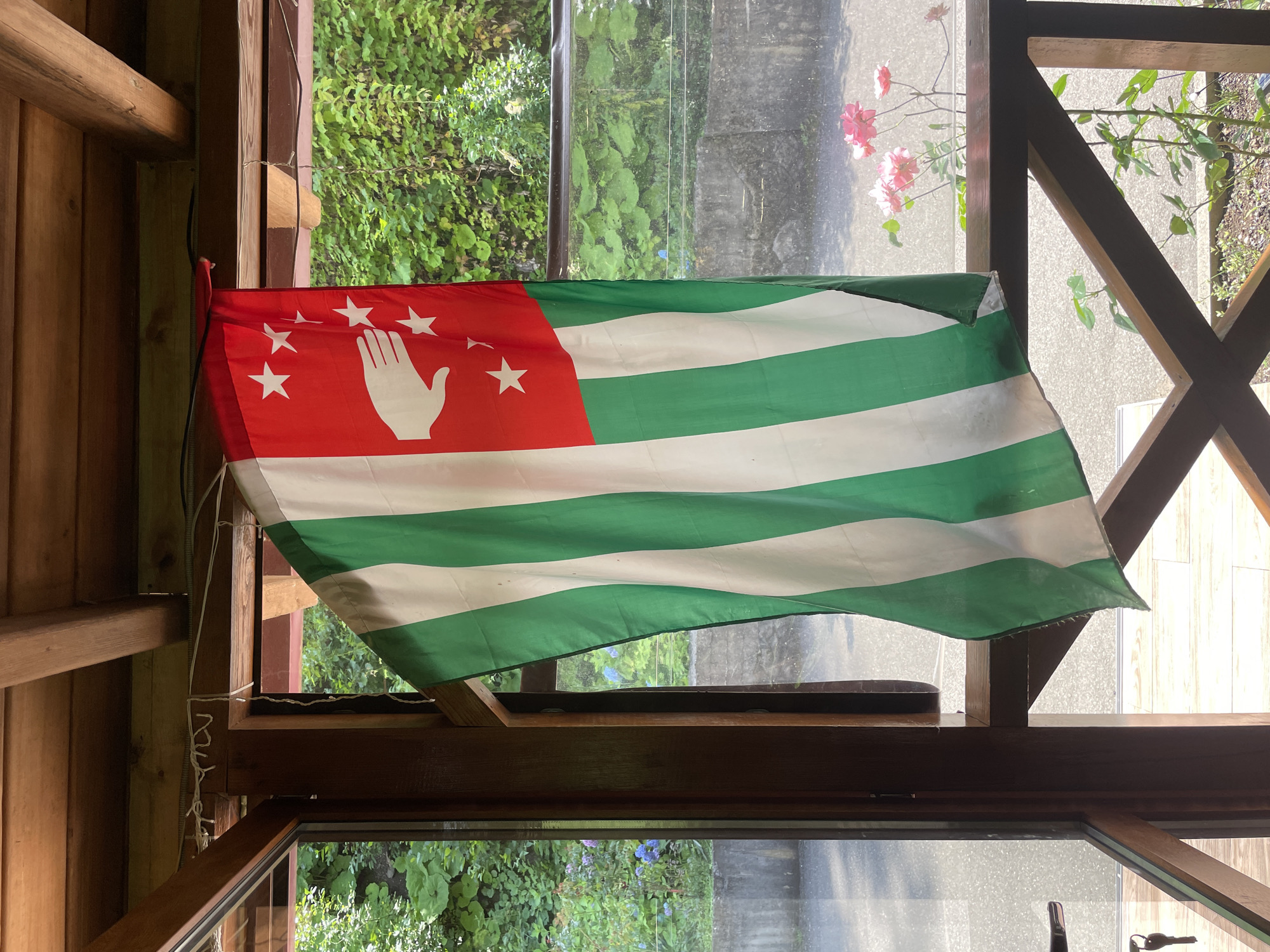A vertical flag, predominantly white and green striped with a red canton, is displayed outdoors, likely from a wooden structure. The red canton features a white open hand symbol and five white five-pointed stars arranged in an arc above and around the hand. The flag is partially obscured by its own folds and the wooden framing of what appears to be a balcony or porch, with a glass window reflecting greenery and some pink flowers in the foreground. The background shows lush green foliage, suggesting a natural or semi-natural setting, possibly within the vicinity of Tqwarchal, Abkhazia. The flag is suspended from above by a white cord.