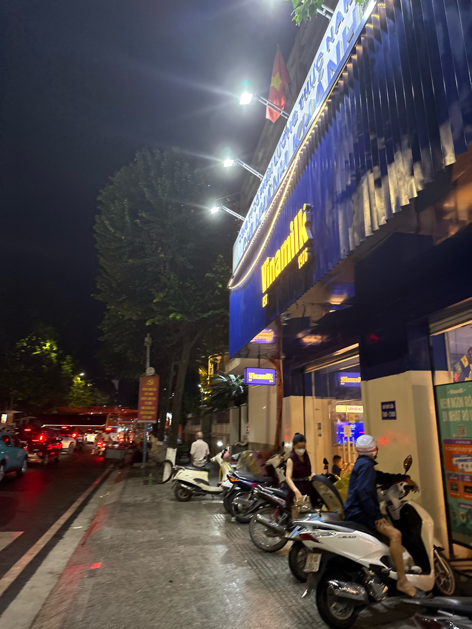 This image captures a nighttime street scene in Nha Trang, Vietnam, near a Vinamilk store. The Vinamilk building features a prominent blue facade with vertical metallic accents and a large, illuminated sign displaying the Vinamilk logo and the year "1976". A Vietnamese flag is visible on a pole attached to the building.
The street is busy with traffic, including cars and numerous motorbikes, a common mode of transportation in Vietnamese cities. Several people are present, some on motorbikes and others standing. One woman is seen sitting on a motorbike, wearing a mask and a dark dress, while a man in a blue jacket and white helmet is also on a motorbike. Another man in a light-colored shirt is seated on a scooter.
The pavement area in front of the store is lined with parked motorbikes, and there are some potted plants. The streetlights cast a bright glow, illuminating the scene and creating reflections on the wet asphalt, suggesting recent rain. A large tree with dense foliage stands to the left of the building, partially obscuring the sky. Signs with Vietnamese text are visible on the building and on a pole on the sidewalk. The overall atmosphere is that of a vibrant urban environment at night.