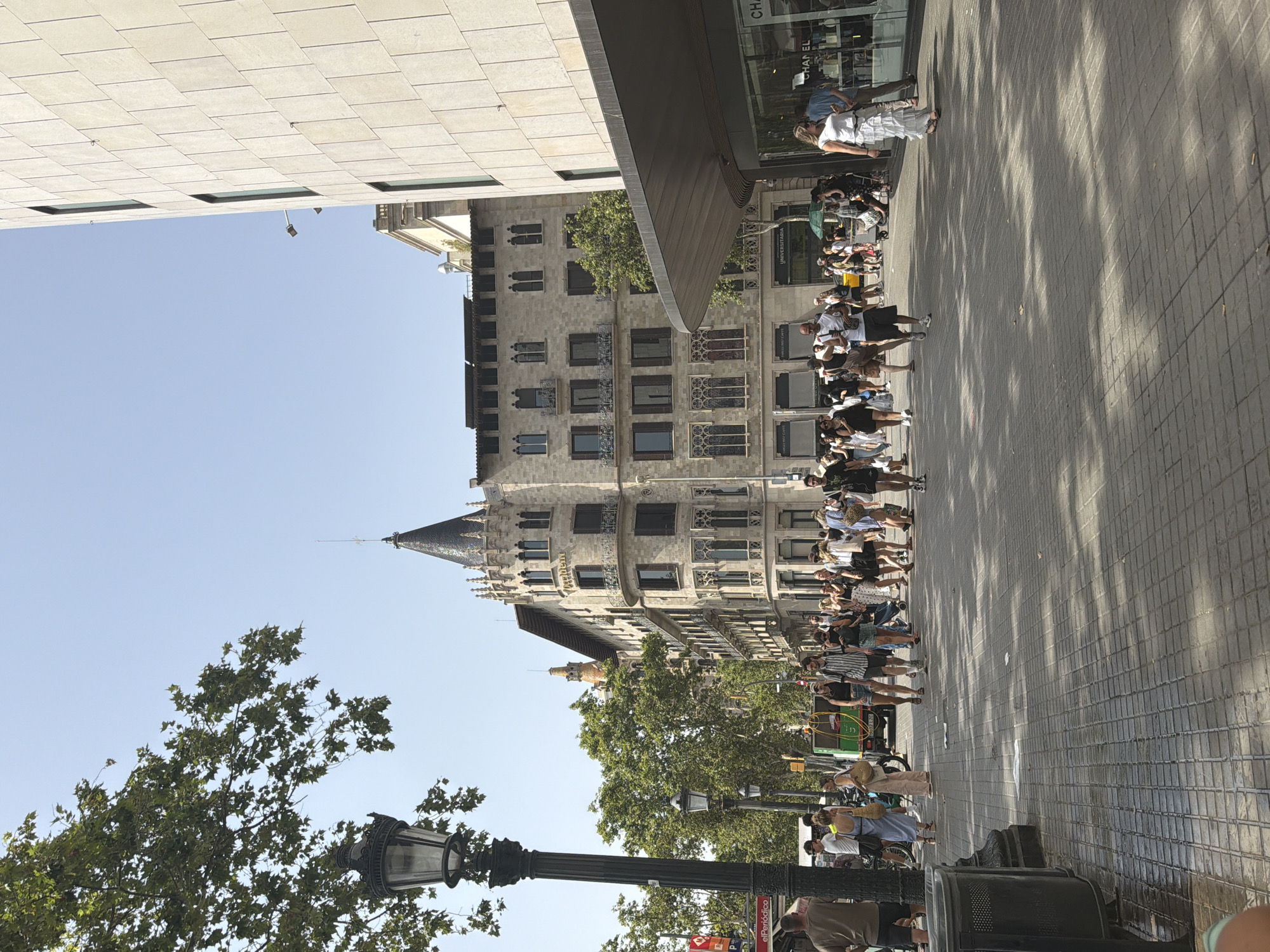 The image captures a street scene in Barcelona, Spain, likely on a sunny day. A diverse group of people are walking on a tiled pedestrian sidewalk, casting shadows from the overhead trees and buildings. The architecture features a prominent ornate building with Gothic Revival elements, including a distinctive spire, alongside a modern building with a smooth, light-colored facade. On the left, a traditional lamppost stands tall. In the background, a green utility vehicle and various shops, including one with "CHANEL" visible, suggest a commercial area. The overall atmosphere is one of a busy, urban environment.