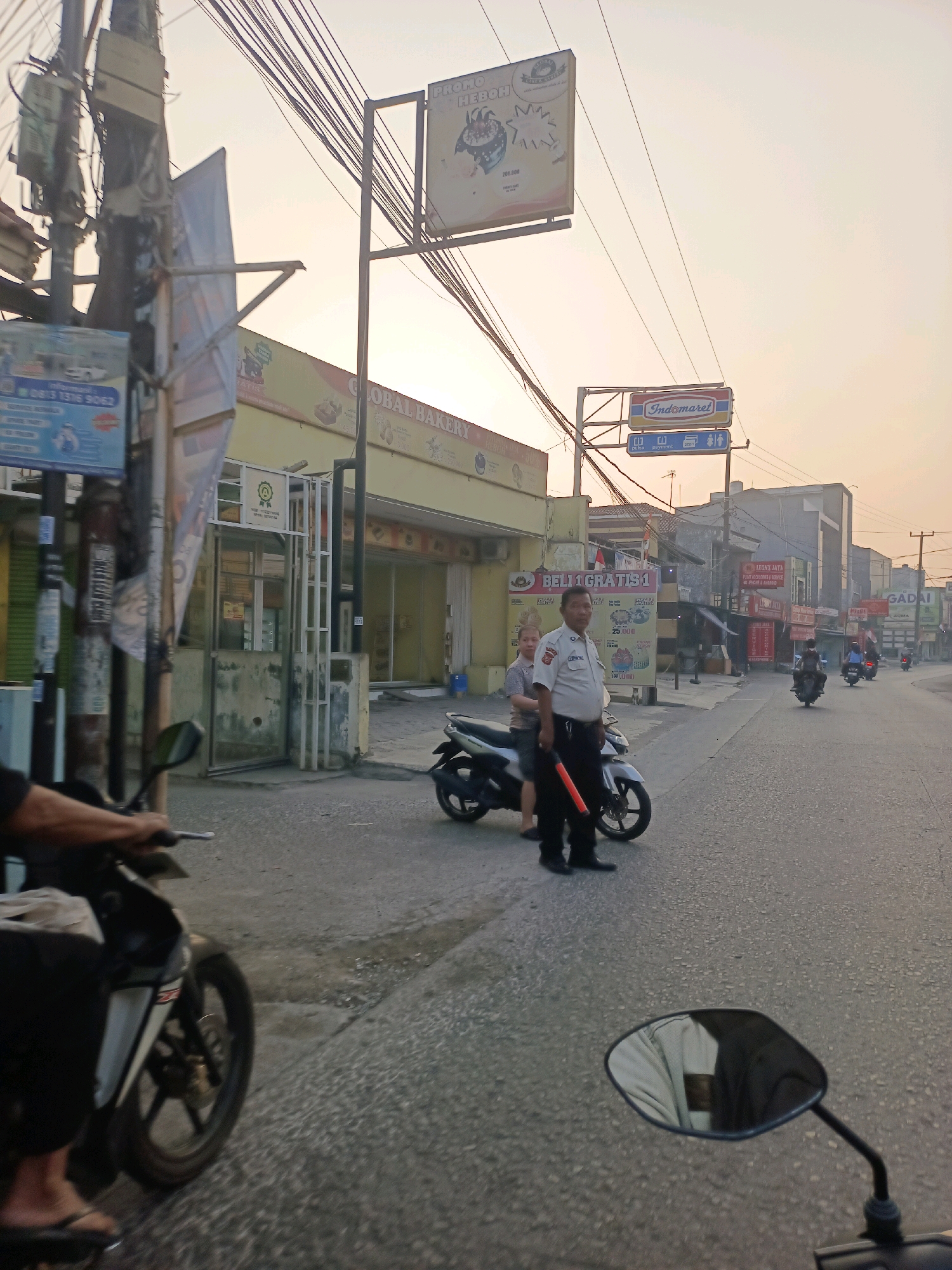 The image depicts a street scene in Karawang, Indonesia, during daylight. A man in a white uniform, possibly a security guard or traffic officer, stands with a woman next to a motorcycle. The man holds a red signaling baton. Behind them, a yellow building with "GLOBAL BAKERY" signage is visible, along with other commercial establishments. A prominent Indomaret sign indicates a nearby convenience store. Several motorcycles are present, with some moving along the road. The sky is overcast with a warm, hazy glow. The street appears to be a typical urban thoroughfare with overhead wires and utility poles.