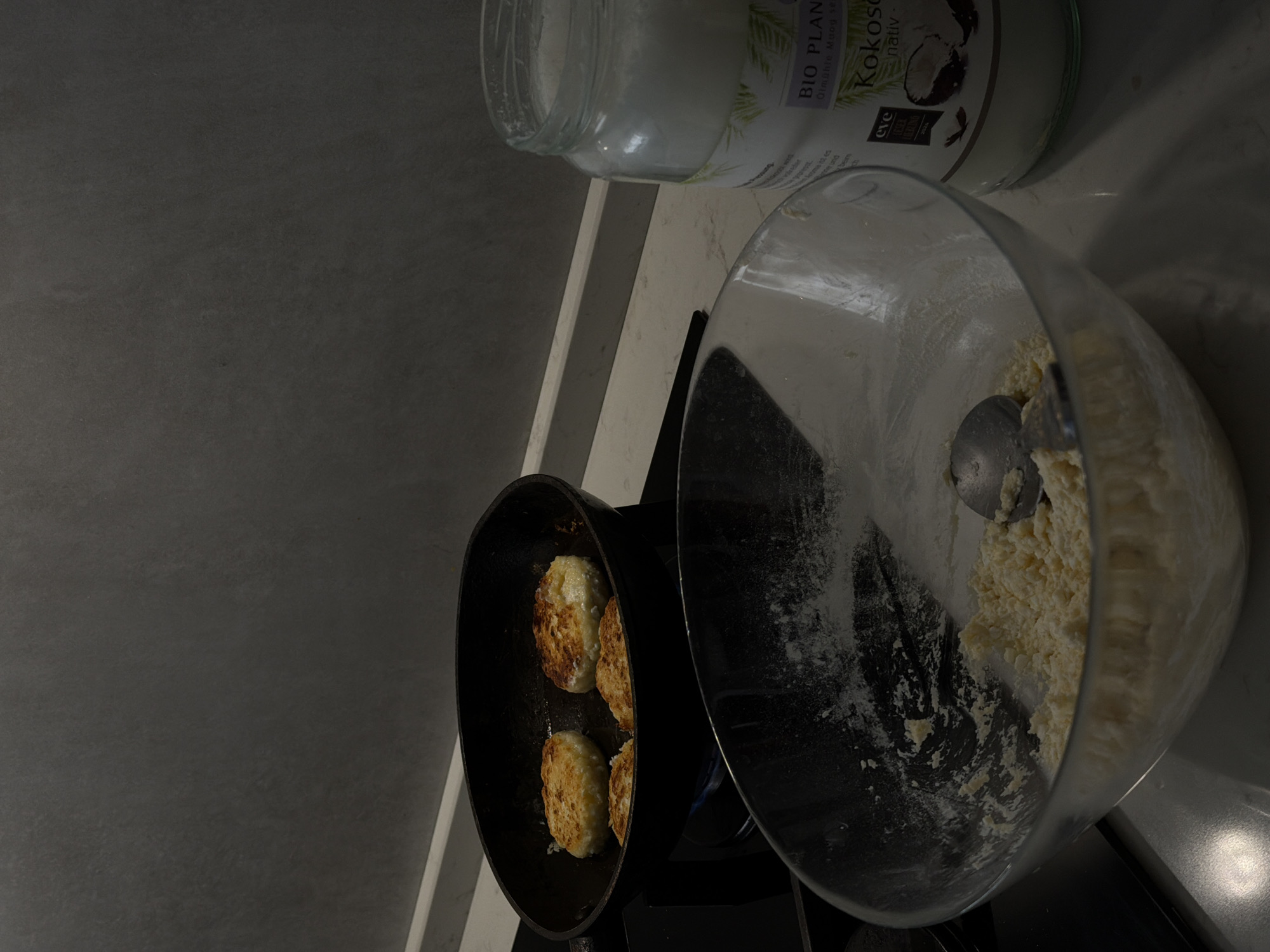 The image captures a kitchen scene in Sofiivska Borshchahivka, Ukraine, during what appears to be evening or night due to the dim lighting. The primary activity is cooking, specifically frying small, round, golden-brown items in a black frying pan on a stovetop. There are three such items, resembling fritters or pancakes (likely syrniki, a Ukrainian cheese pancake), with visibly crispy, browned surfaces. A faint blue flame indicates the burner is active.
To the right of the frying pan, a large, clear glass mixing bowl holds a light-colored, lumpy mixture, consistent with dough or cheese preparation, with flour residue visible on the inner walls. A metal spoon rests within this mixture.
In the upper right background, a glass jar of "BIO PLANT Kokosöl nativ" (native coconut oil) is visible, with a white label featuring green leaves and the "EVE VEGAN" certification logo. The oil inside appears solid and white, typical of coconut oil at room temperature. The items are arranged on a light-colored countertop. The overall setting suggests a domestic kitchen environment focused on meal preparation.
