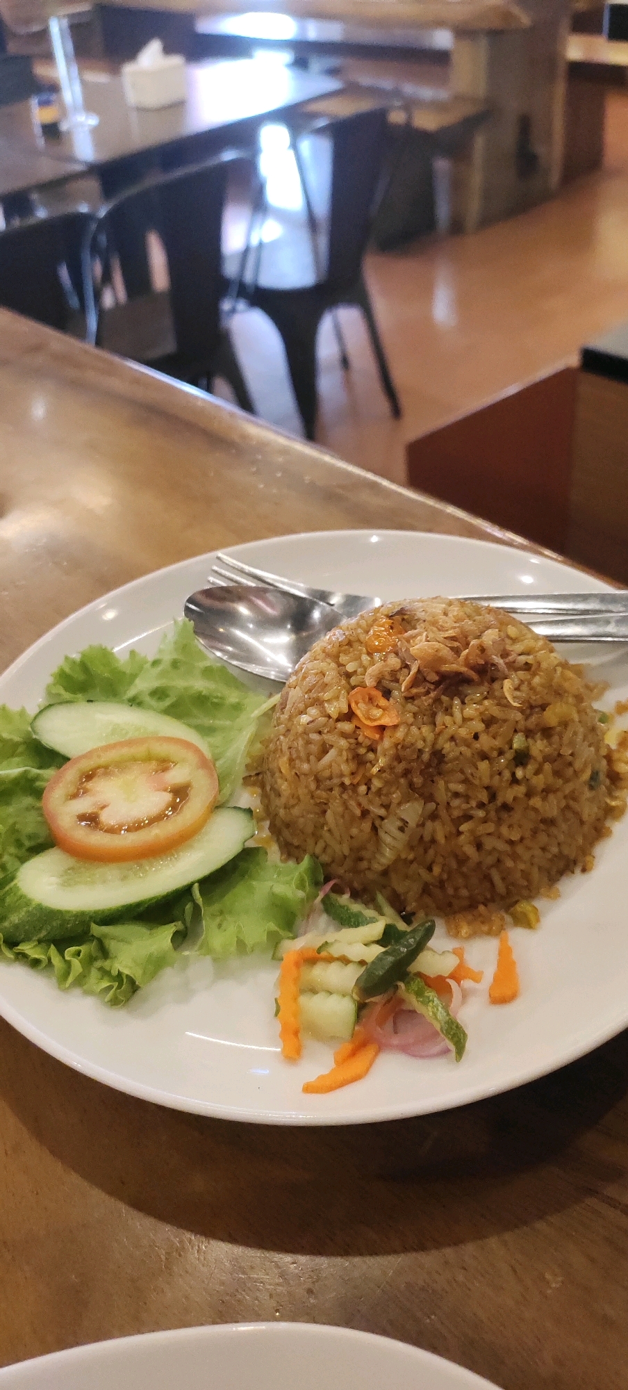 An overhead, medium shot captures a white plate of Indonesian fried rice on a wooden table in a restaurant. The fried rice is piled high, topped with crispy fried shallots and small pieces of chili and shrimp. To the left of the rice are fresh lettuce leaves, two slices of cucumber, and one slice of tomato. In the bottom right corner of the plate is a small portion of pickled vegetables, consisting of thinly sliced carrots, cucumbers, red onions, and a green chili. A spoon and fork are resting on the edge of the plate. The background is blurred, showing tables and chairs typical of a casual dining establishment. This scene is located in the city of Nganguk, Indonesia.