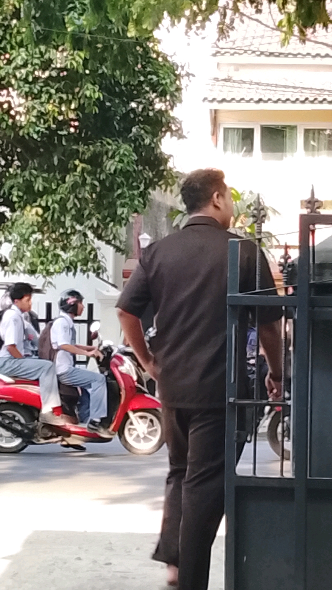 A man in a dark short-sleeved shirt and dark pants stands with his back to the camera, partially obscured by a dark metal gate. In the background, two young males wearing school uniforms and helmets are riding a red scooter. The scooter has a white front wheel. Behind the scooter, a building with a tiled roof and white window frames is visible. Green foliage from a tree hangs over the scene. The pavement in the foreground is a light color.