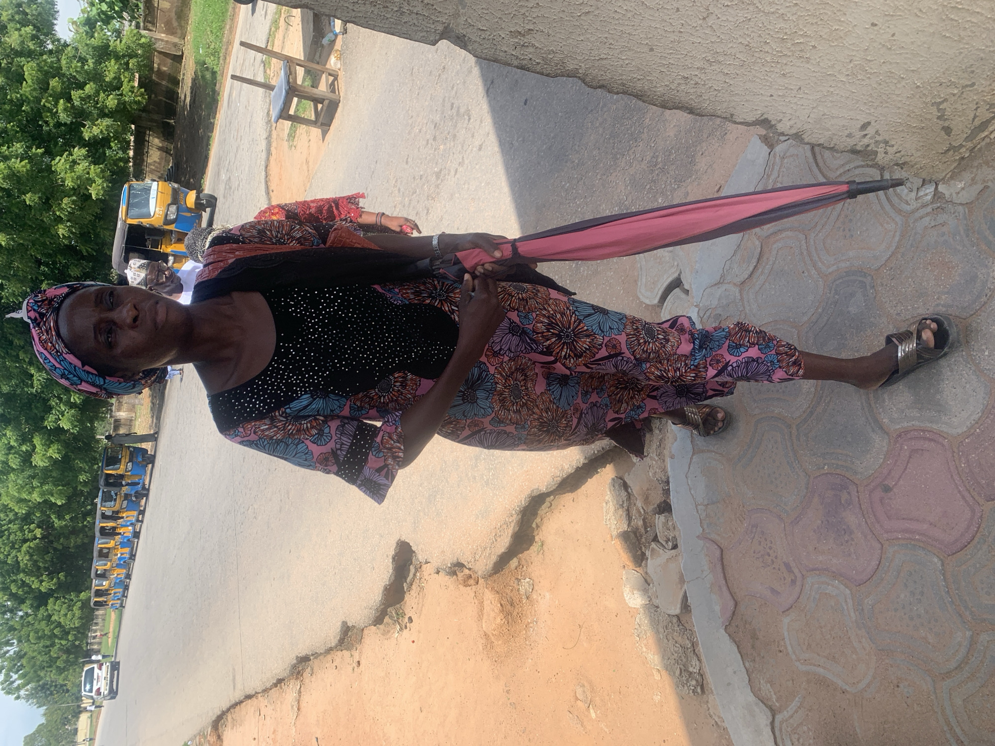 The image shows a woman standing outdoors, likely in Jalingo, Nigeria, given the context. She is facing the camera and appears to be looking slightly to her left. She is wearing a black top with embellishments and a brightly patterned skirt with floral designs in pink, blue, and orange. A colorful headscarf, predominantly pink with blue and black accents, adorns her head. She is also holding a partially open pink umbrella with a black handle. Her left hand is visible, adorned with a bracelet.
The woman is standing on a paved area made of interlocking bricks. To her left, a dirt path or road is visible, with a concrete curb separating it from the paved area. In the background, several bright yellow and blue auto-rickshaws (also known as "keke") are parked in a line on a road. Further back, lush green trees line the area, suggesting a warm, possibly sunny day. The lighting and shadows indicate it is daytime. A small white van is parked on the road in the distance, and a wooden chair with a blue seat is visible near some greenery. The overall impression is a street scene in a populated area, likely in a market or transport hub. The woman's expression is neutral. No visible text is present in the image.