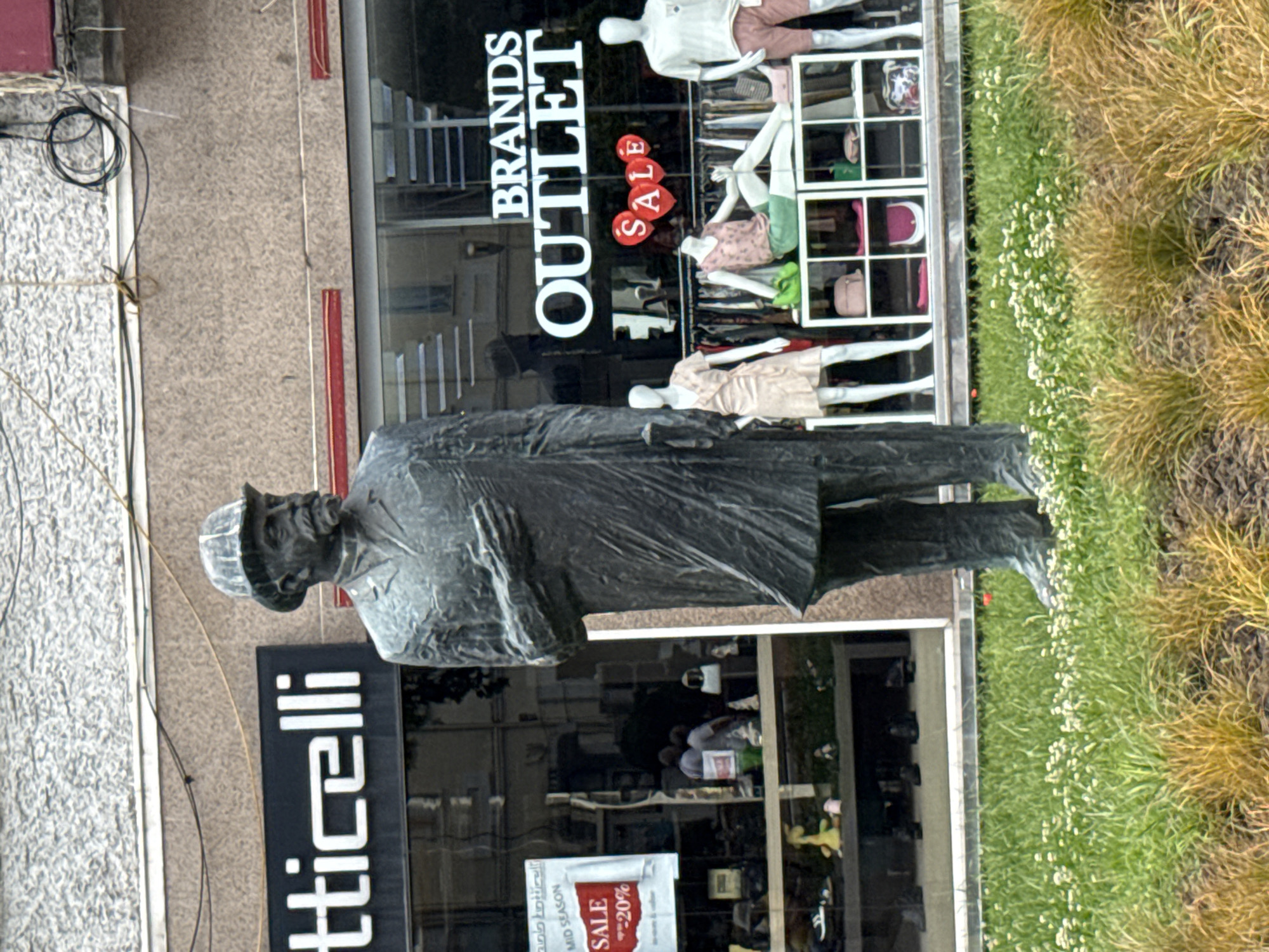A full-body, outdoor, eye-level, medium shot captures a bronze statue of a man in a suit and hat standing in front of a storefront. The man is facing right, with his left hand on his chest and his right hand holding a cane. The storefront is a retail establishment with large glass windows displaying mannequins in clothing. The words "BRANDS OUTLET" are visible in large white letters on the window. Below this, red heart shapes with the word "SALE" are displayed. In the reflection of the window, two people are visible, appearing to be indoors. To the left of the statue, a smaller sign indicates a sale with "-20%". The ground in front of the storefront is covered in green grass and some yellowing foliage. The building facade above the storefront is textured and appears to be made of concrete. Overhead, electrical wires are visible against the building.