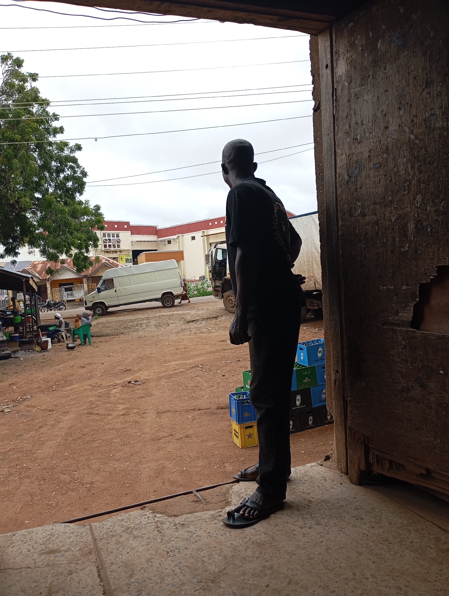 The image depicts an outdoor scene in what appears to be a market or roadside area in Shendam, Nigeria. In the foreground, a man stands facing away from the camera, wearing a black t-shirt and black pants. He is wearing black sandals. To the man's right, there is a wooden door or entryway with a weathered and damaged surface. Behind the man, a dirt ground is visible, leading to various structures.
Further in the background, there are several elements suggesting a commercial or transportation hub. A white van is parked on the right side of the road. To the left of the van, a building with a red and white facade is visible. There are stacks of crates, some blue and green, with labels that appear to be for beverages. In the distance, a large truck is parked. On the far left, there is a tree with green foliage, and further left, more stalls or structures are partially visible, along with a motorcycle. Two individuals are seated on chairs in the shaded area under one of the stalls, and cooking pots are also present. The sky is overcast.
