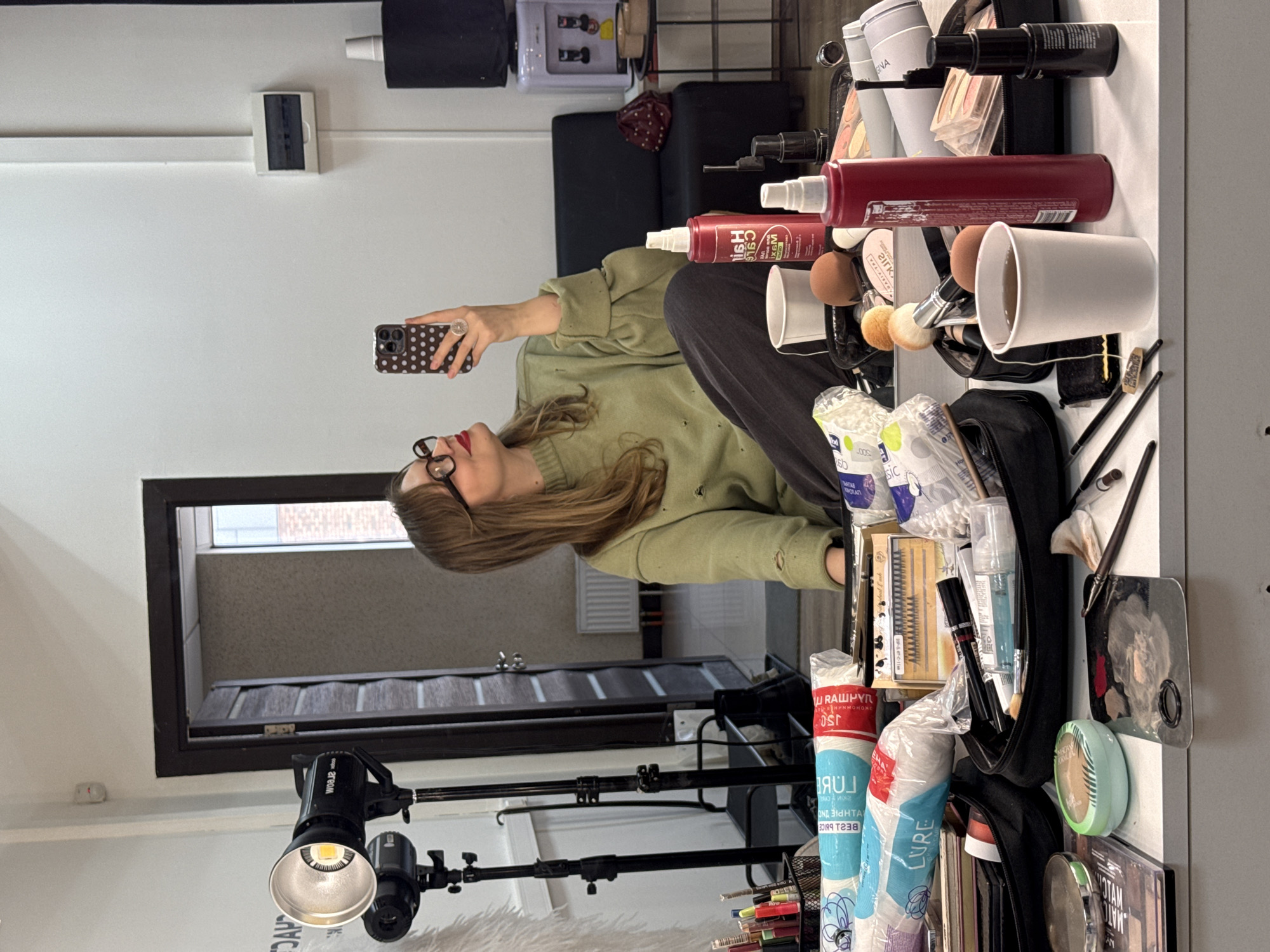 Here is a detailed description of the image:
**General Description:**
The image shows a woman in a casual outfit taking a selfie in what appears to be a beauty studio or salon setting. The studio has makeup products and equipment visible on a table. The image is slightly rotated clockwise.
**People:**
* A woman with blonde/light brown hair, wearing glasses, a green long-sleeved shirt, and dark pants, is taking a selfie with her phone. She has red lipstick on.
**Subjects:**
* Makeup products and tools are prominently displayed on a white surface.
* A professional photography light with adjustable arm is positioned in the foreground.
* A mirror is visible, reflecting a view of what seems to be the room's entrance.
**Objects:**
* Several makeup brushes and sponges, a variety of makeup containers (bottles, tubes, compacts), and cotton swabs.
* A black photography light with stand.
* A black makeup bag.
* Hair products in red and white bottles.
* An iPhone with a dotted case.
* A door or large window with a black frame.
* A white wall with light switches and other fixtures.
* A black cushioned chair in the background.
**Actions:**
* The woman is holding her phone and taking a selfie.
**Interactions:**
* The main interaction is the woman using her phone to capture the selfie in the studio setting.
**Environment:**
* The setting is a beauty studio or salon, with a clean and well-lit area.
* The wall is painted white.
* The room has a window or door leading to another area.
**Location:**
While I cannot confirm the exact location in Izhevsk, Russia, based solely on this image, the setting is likely a beauty salon, makeup studio, or a similar establishment.