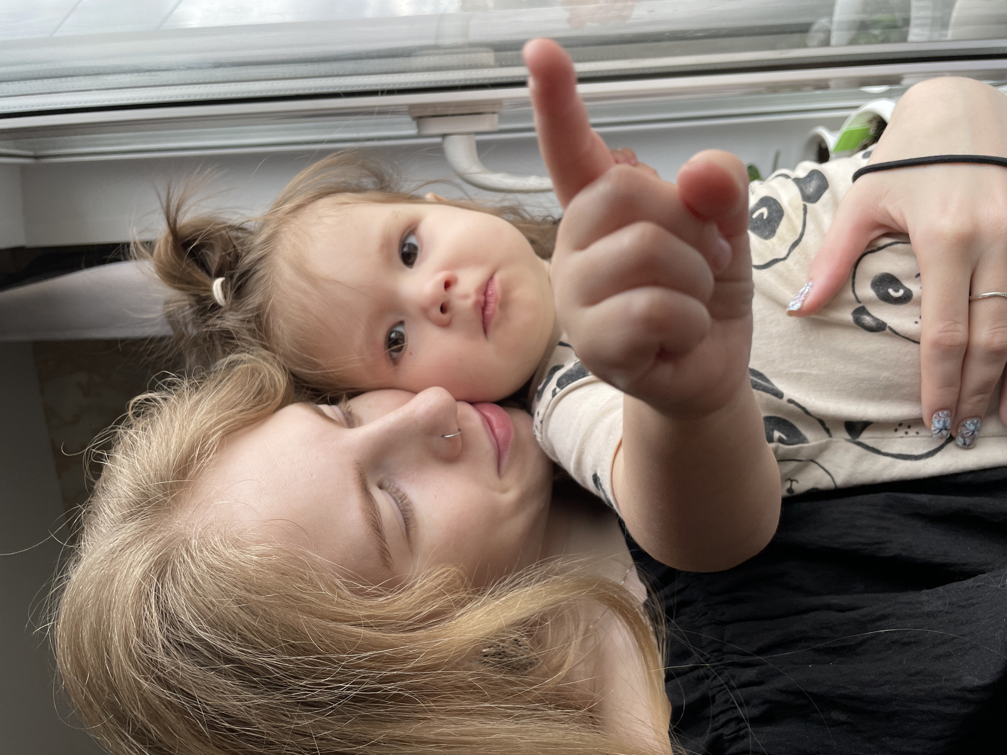 An intimate portrait captures a young woman and a small child lying together, possibly a mother and daughter, taken indoors next to a window in Izhevsk, Russia. The image is oriented vertically, showing the subjects from an overhead perspective, with the window frame visible above them.
The young woman, likely the mother, occupies the lower and central part of the frame. She has long, wavy blonde hair that cascades around her face and shoulders. Her eyes are closed, and a gentle smile plays on her lips, suggesting a moment of peace or affection. She wears a silver nose ring and a subtle silver ring on a finger of her visible right hand. A black elastic hair tie is wrapped around her right wrist, and she is dressed in a dark, possibly black, top.
Resting her head close to the woman's cheek, the small child looks directly at the camera with wide, dark eyes and a serious, inquisitive expression. She has light brown hair, pulled back into a small, spiky bun on top of her head. The child is wearing a light-colored t-shirt adorned with outlines of black panda bears. Her right hand is raised, index finger pointing outwards towards the viewer, adding a playful and engaging element to the scene. Her fingernails appear to have a light polish or glittery pattern.
The setting appears to be a cozy indoor space, illuminated by natural daylight filtering through the large window that runs along the top of the frame. Through the window, faint hints of greenery or outdoor foliage are visible, though the external weather is not clearly discernible beyond being bright. The overall mood of the image is one of tender connection and candid interaction between the two individuals. There is no visible text in the image.