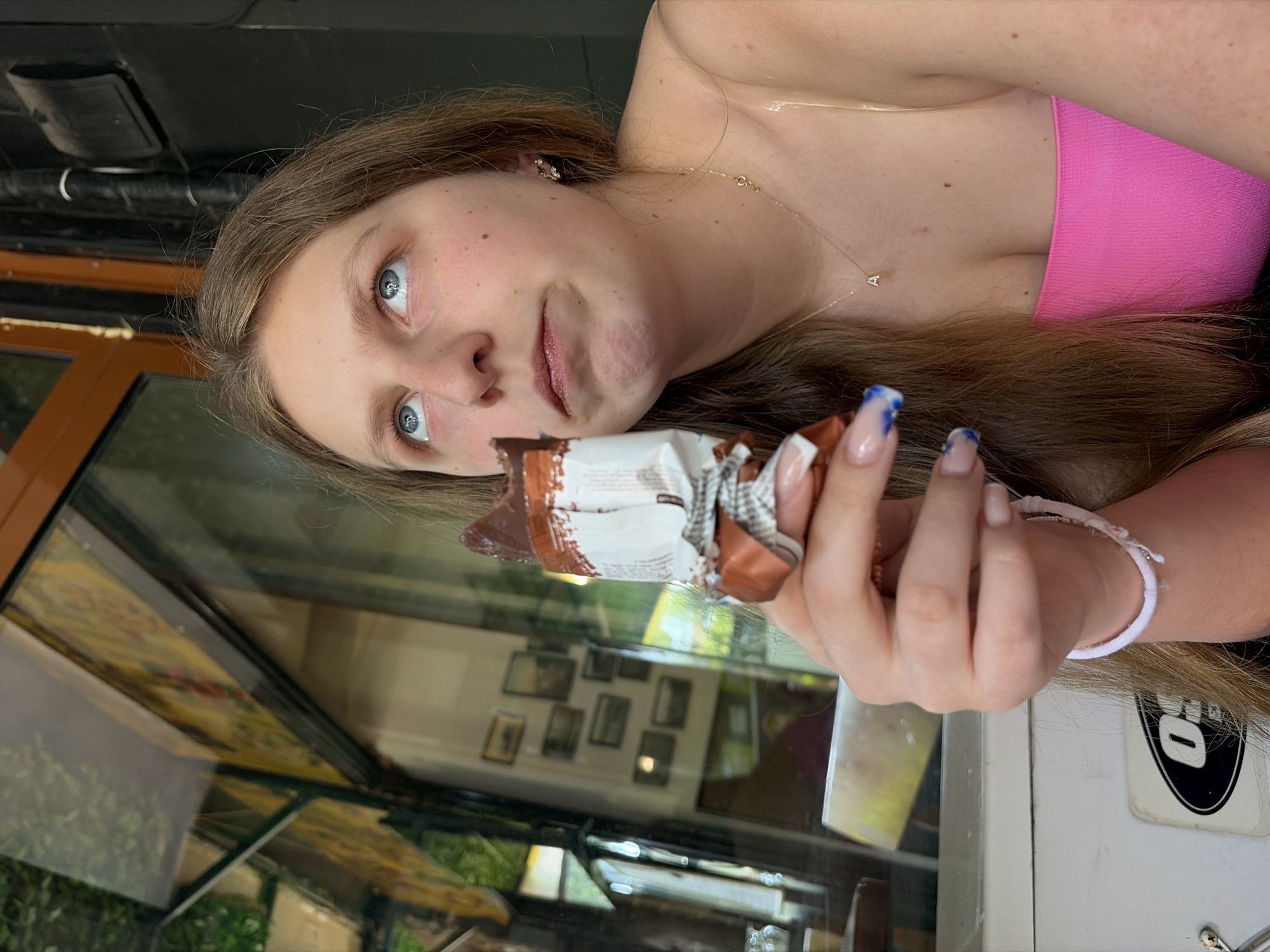 A young woman with long, light brown hair, striking blue eyes, and a fair complexion with freckles is captured in a close-up, rotated image. She is wearing a bright pink strapless top and a delicate gold necklace with a small 'A' pendant. Her expression is playful and slightly mischievous, with wide eyes and a subtle smirk.
In her right hand, she holds a partially unwrapped chocolate bar or similar snack, with visible text on the wrapper including fragments like "WHITE" and "SUGAR." Her fingernails feature a French manicure with some nails adorned with blue and white marbled designs. A white elastic band or bracelet is visible on her left wrist.
The setting appears to be an indoor or semi-enclosed space, possibly a cafe or restaurant, during daytime. A glass surface in front of her reflects greenery from outside, as well as a collection of framed pictures hanging on a distant wall. In the upper background, dark, possibly metallic, structures are visible. A black and white sticker, featuring a prominent 'O' or 'D' within an oval, is affixed to a white surface in the lower right portion of the image.
The woman is engaged in the casual activity of enjoying a snack, seemingly posing for the camera with a lighthearted demeanor. No direct visual cues confirm the specified location of Burgas, Bulgaria, but this is the provided context for the scene.