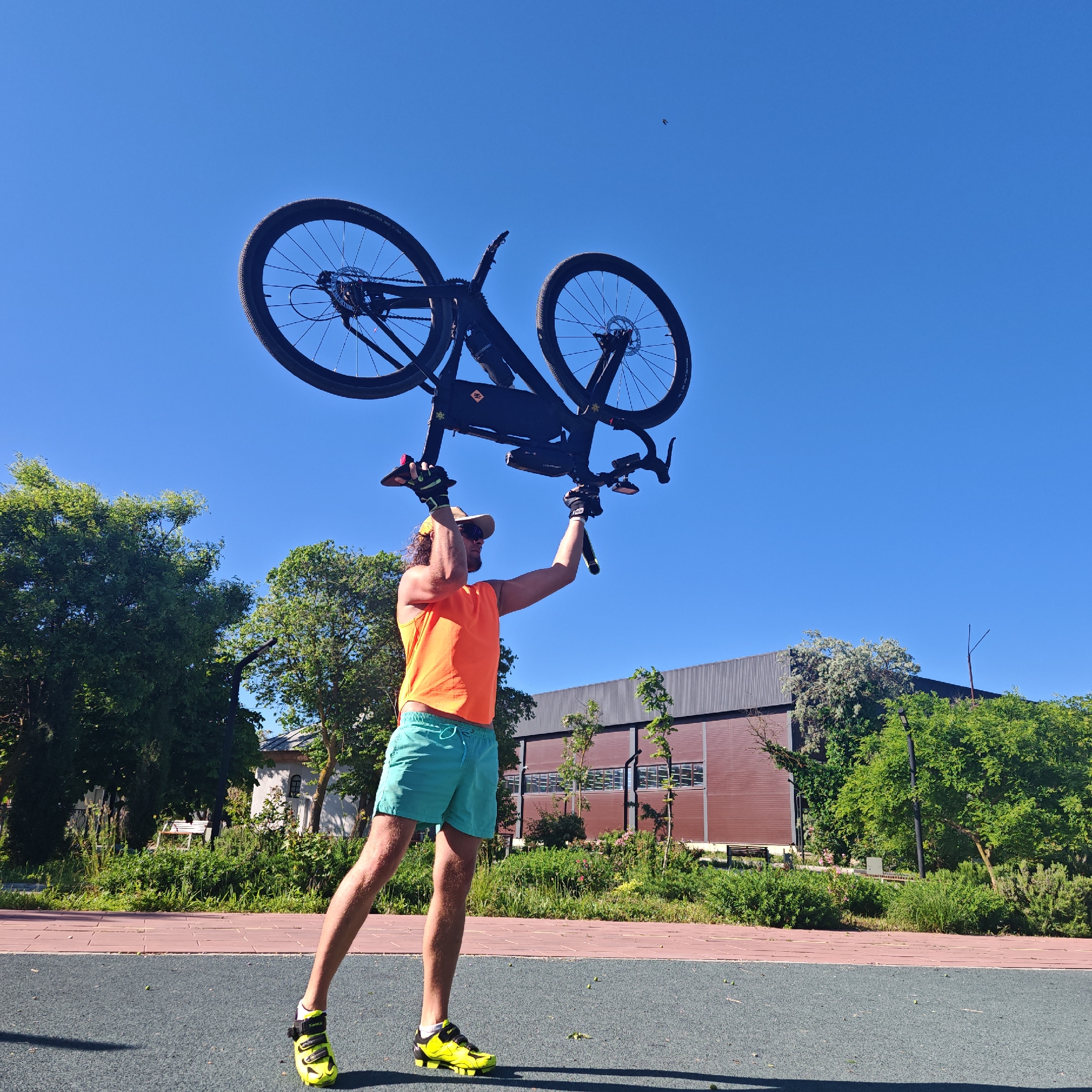 A person is holding a bicycle up in the air. The person is wearing an orange tank top, green shorts, a hat, sunglasses, gloves and yellow and black athletic shoes. The bicycle is black. The location appears to be a park or public space, possibly in городское поселение Щелкино, Ukraine, due to the request, as suggested by the presence of greenery, walkways, and a building in the background. The sky is clear and blue. The subject is lifting the bicycle overhead with both arms extended.
