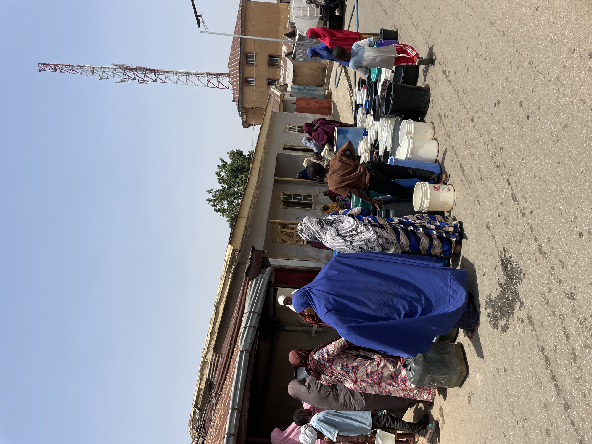 In Jos, Nigeria, a line of people stands along a building's side, their shadows cast on the street. The queue stretches toward a water truck visible in the background, its presence underscored by an array of buckets of various sizes and colors – white, black, and blue – lining the building. People in the line are dressed in traditional clothing, including hijabs and robes of various colors such as blue, red, purple, and patterned fabrics. Their actions suggest a communal effort to collect water. Overhead, a communications tower looms in the distance, contrasting with the building's aged façade. The environment is arid, and the street bears signs of wear.