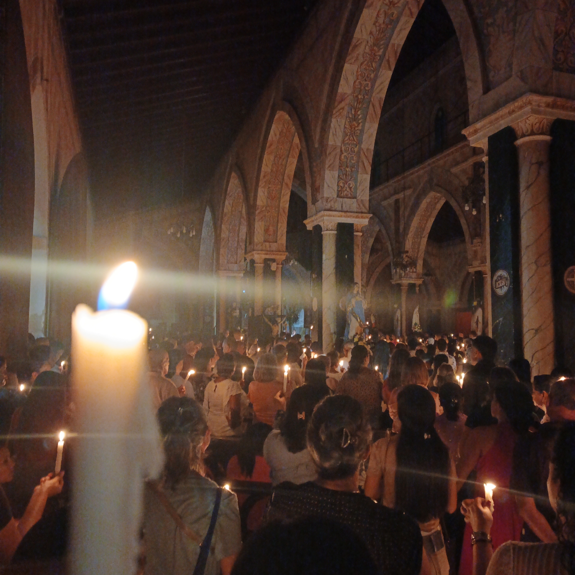 The image shows a crowd of people inside a church holding lit candles. The scene is in a church with archways and columns, decorated with patterns. Statues are visible in the background. The location is Tinaquillo, Venezuela.