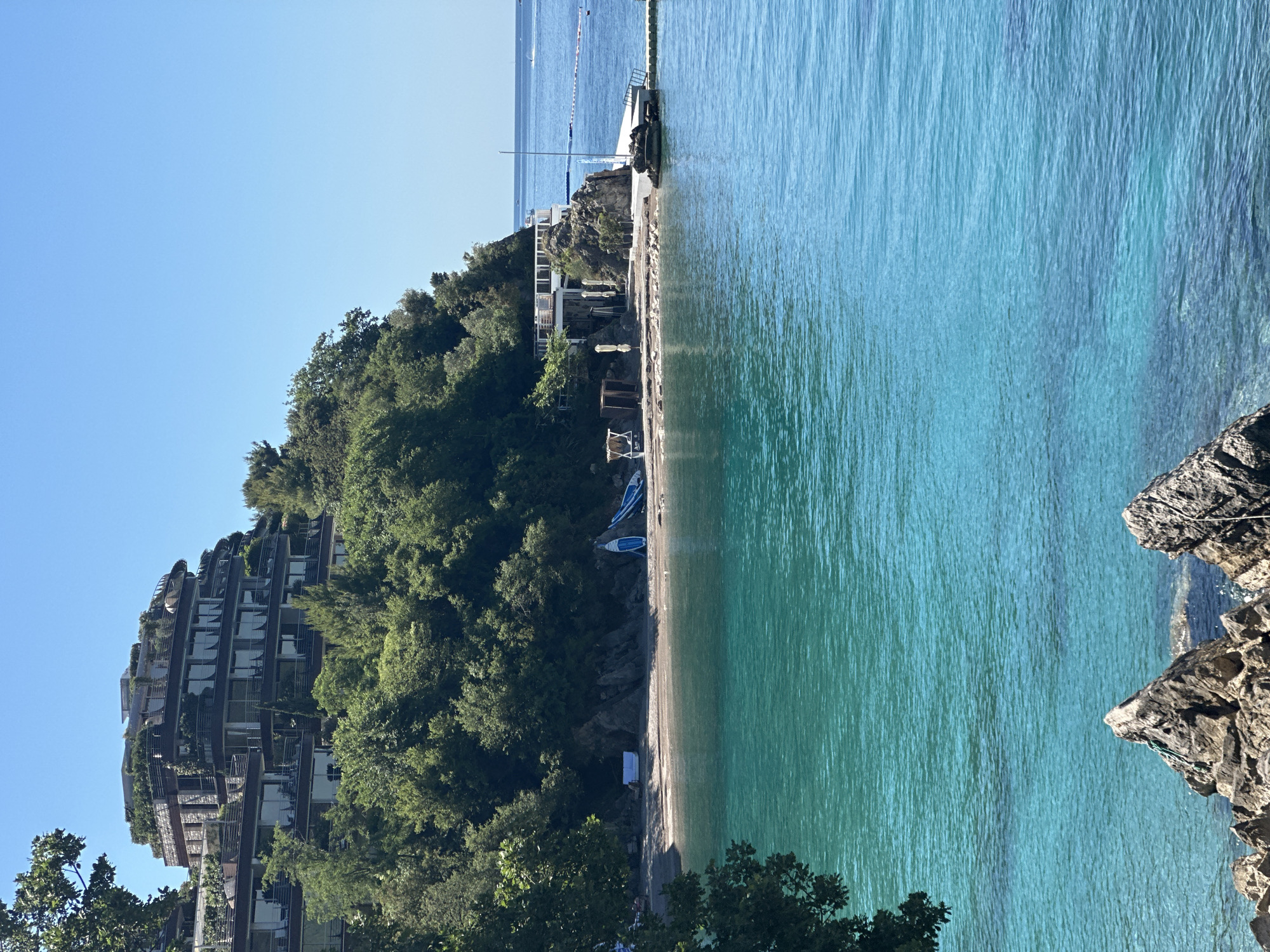 The image shows a coastal scene in Budva, Montenegro. The turquoise water of the Adriatic Sea occupies a significant portion of the frame. A narrow sandy beach is bordered by dense green trees and vegetation that cover a hillside. On the left side of the image, a modern, multi-story building is partially obscured by the foliage. On the beach, there are some small objects, possibly boats. At the far end of the beach, a small pier extends into the water with a structure at its end. In the foreground, rocky formations jut into the sea. The sky is clear and blue.