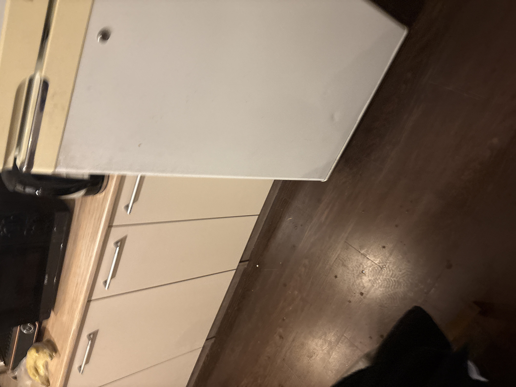 A close-up, angled shot shows a section of a kitchen with a refrigerator, cabinets, and a wooden floor. The refrigerator is white with a cream-colored top, and appears to be a retro model. The kitchen cabinets are a light beige color with horizontal silver handles. On the countertop next to the refrigerator, a peeled banana is visible. The wooden floor is dark brown and appears somewhat worn, with some small debris scattered on it. The scene is indoors, likely in a residential kitchen. No people are visible in the image. The location in the city of Orekhovka, Russia is not discernible from the visual information provided.