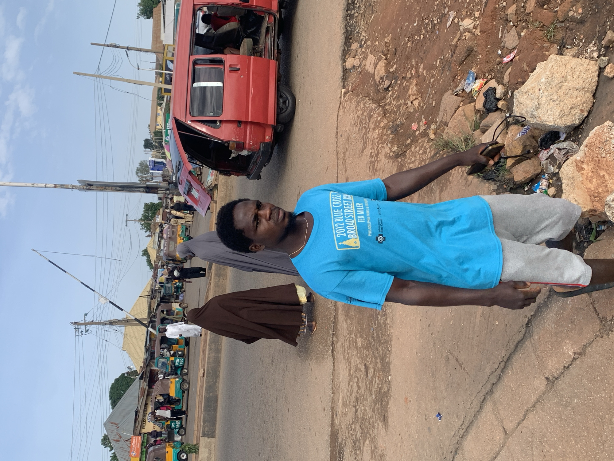 In Gura Top, Nigeria, a man in a blue t-shirt and grey shorts stands on a dirt road with scattered debris. Behind him, a red van with its rear and side doors open is parked, with passengers visible inside. Several auto-rickshaws are lined up on the street. In the background, buildings with corrugated metal roofs are visible, along with a utility pole and power lines. Two women in traditional attire, one in a brown hijab and the other in a grey hijab, walk in the middle ground. A white striped barrier is raised above the road. The sky is clear and bright.