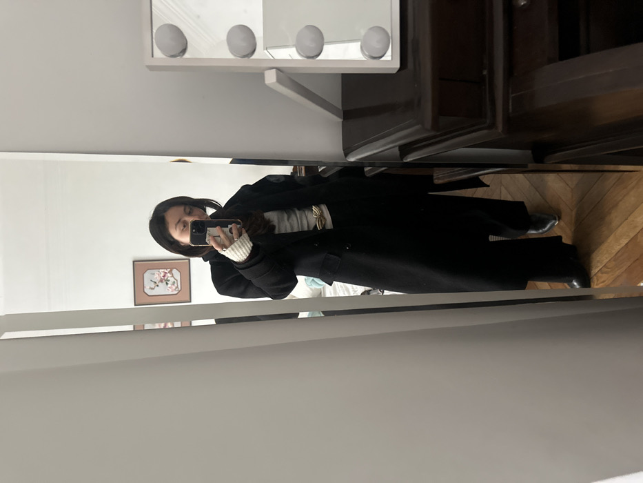This image depicts a woman taking a mirror selfie indoors. The woman, with dark hair, holds a smartphone in her right hand, positioned in front of her face, partially obscuring her features. She is dressed in a black double-breasted overcoat, a light-colored long-sleeved top, black wide-leg trousers, and black pointed-toe boots. A golden-colored, stylized belt buckle is visible at her waist, and a light-colored knitted wrist warmer is on her left wrist, adorned with rings. The phone case features multiple decorative stickers. The reflected indoor environment reveals light wooden parquet flooring, white walls, and a framed piece of artwork with floral motifs on the wall behind the subject. To the right of the mirror, a dark wooden dresser or vanity table supports a white vanity mirror equipped with four visible circular LED bulbs. The interior architectural style, particularly the wooden parquet flooring, is consistent with residential buildings found in the city of Paris, France.