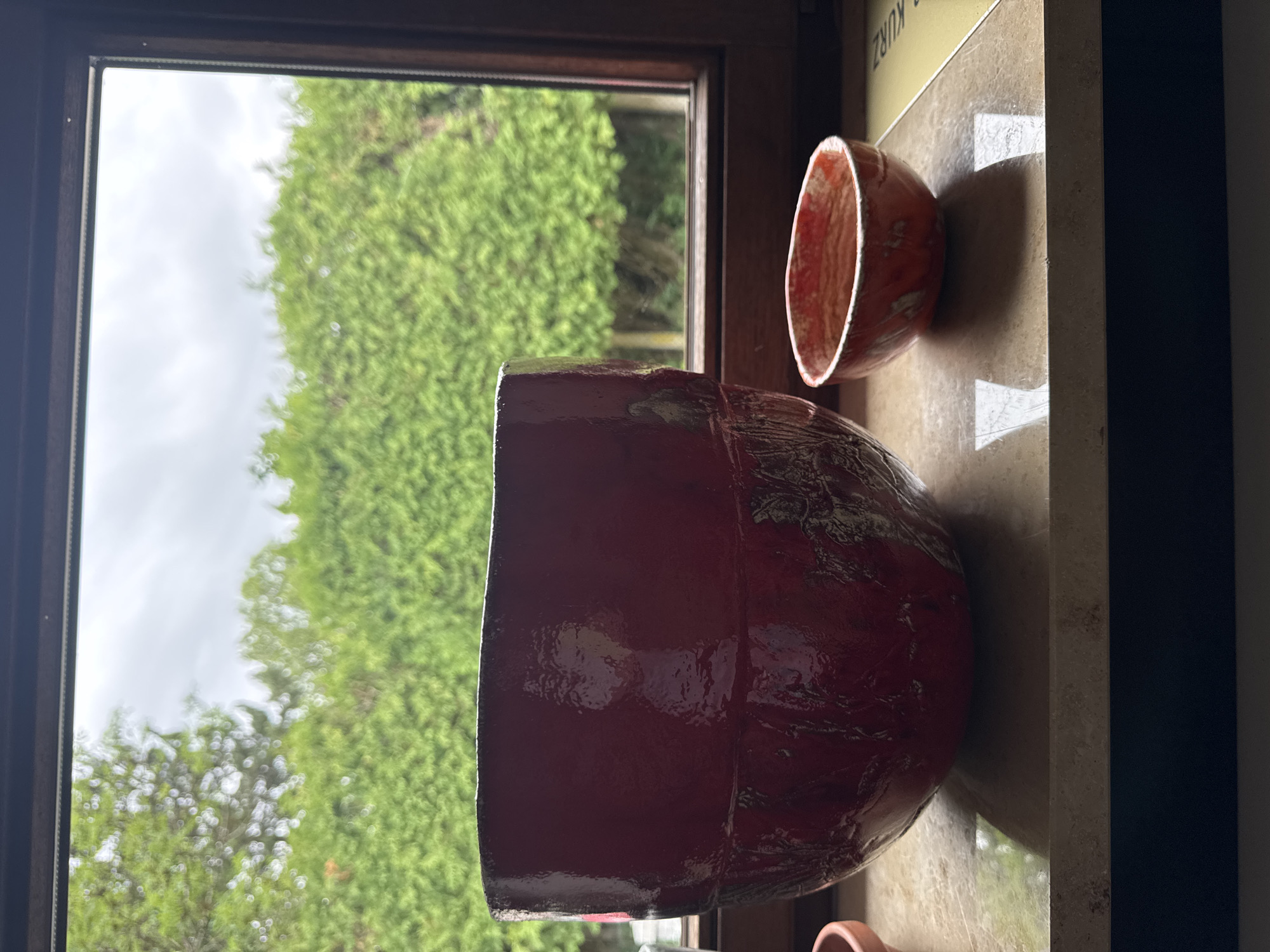 The image shows two ceramic pots on a windowsill. The larger pot is dark red with a textured surface. The smaller pot is orange with a lighter colored interior and a similar textured exterior. The pots are placed on a speckled countertop. In the background, through a window, is a dense green hedge and a cloudy sky. The scene is indoors, likely in a residential setting, as suggested by the windowsill and view of a garden hedge. There are no people or animals present. The lighting suggests it is daytime, but overcast, with the light coming from the window. The setting appears to be a home in Hohenbrunn, Germany.