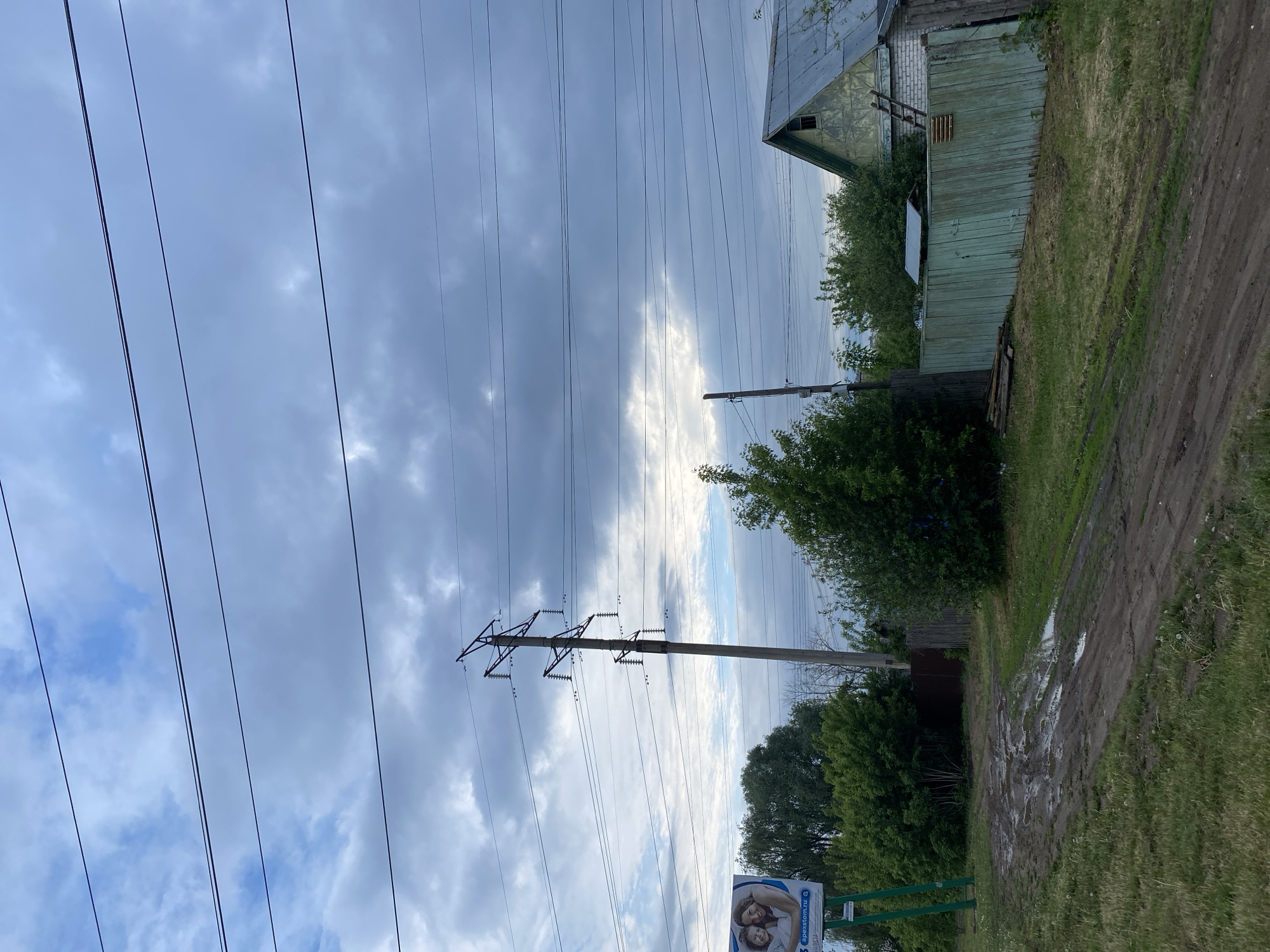 The image presents a rural or semi-rural scene, likely in Chaikovsky, Russia, based on potential signage information. A dirt road, heavily muddied, runs alongside a grassy verge. A weathered wooden building with a corrugated metal roof stands to the right of the road. Power lines and utility poles stretch across a cloudy sky. Vegetation, including trees and bushes, is scattered throughout the scene. A billboard or sign with a partial image of people is visible in the lower portion of the image.