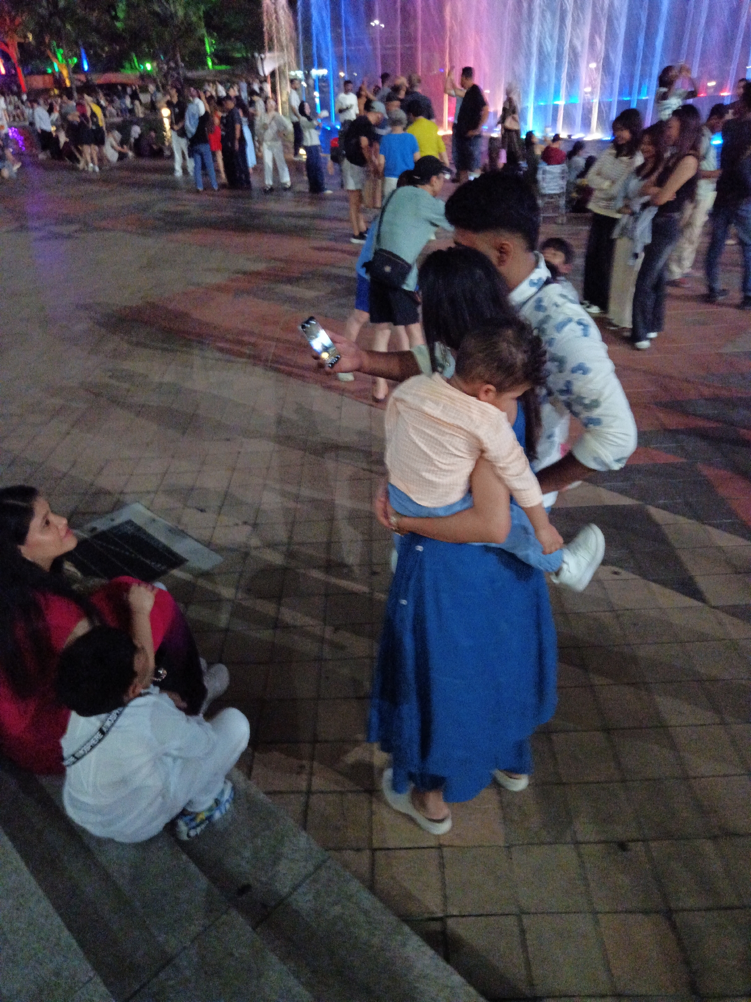 This image depicts a nighttime scene at the Symphony Lake water fountain show in Kuala Lumpur City Centre Park, Malaysia. In the foreground, a woman in a red top and dark pants is seated on steps, looking to her left. Beside her, a child in a white outfit with a black strap is also seated on the steps, facing away from the camera. In the mid-ground, a person in a blue dress is holding a young child wearing a peach-colored shirt and blue pants. Another adult, wearing a patterned shirt, stands behind them. The background features a crowd of people gathered around a colorful water fountain display. The lighting is a mix of natural darkness and artificial illumination from the fountain and surrounding lights.