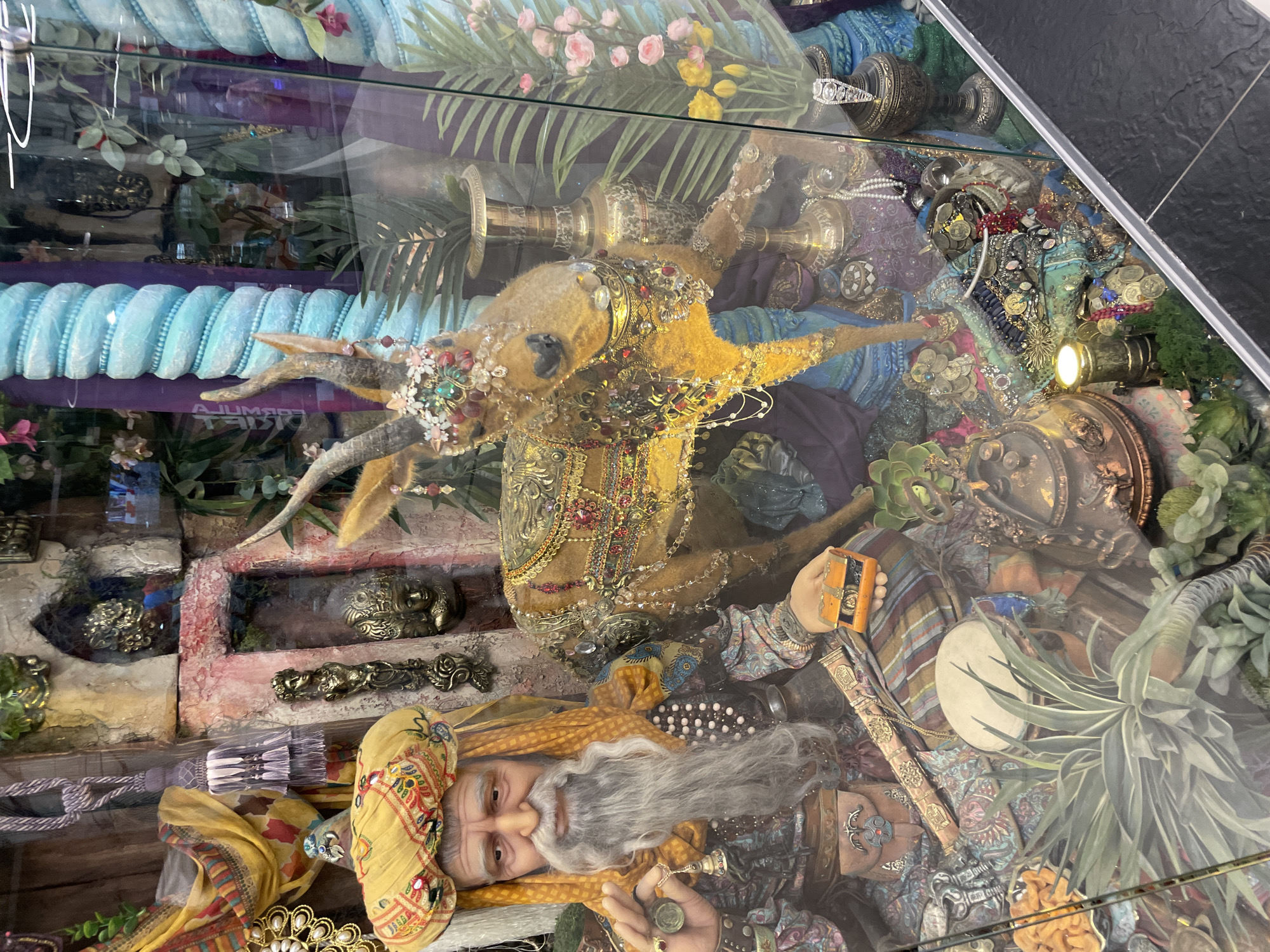 The image shows a diorama display inside a glass case. It depicts a fantastical, possibly Middle-Eastern themed scene. At the bottom of the display is a human figure with a long, gray beard, wearing a turban and ornate clothing. A jeweled ram figure lies diagonally across the center, adorned with jewelry and placed among various brass and gold ornaments. At the top of the display, there are artificial flowers and plants. The overall scene gives the impression of a treasure trove. The location is unspecified.