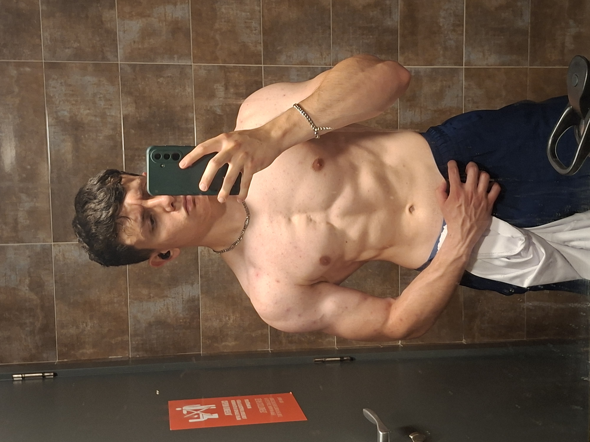 A shirtless young man is taking a selfie in a restroom. He is holding a dark green smartphone. He has a silver chain necklace and a bracelet on his left wrist. He is wearing dark blue pants and a white shirt is partially visible near his hip. The restroom has brown tiled walls and a dark gray door. There is a red sign with white text and a picture of a person in a wheelchair on the door. The exact location in Plovdiv, Bulgaria cannot be determined from the image.