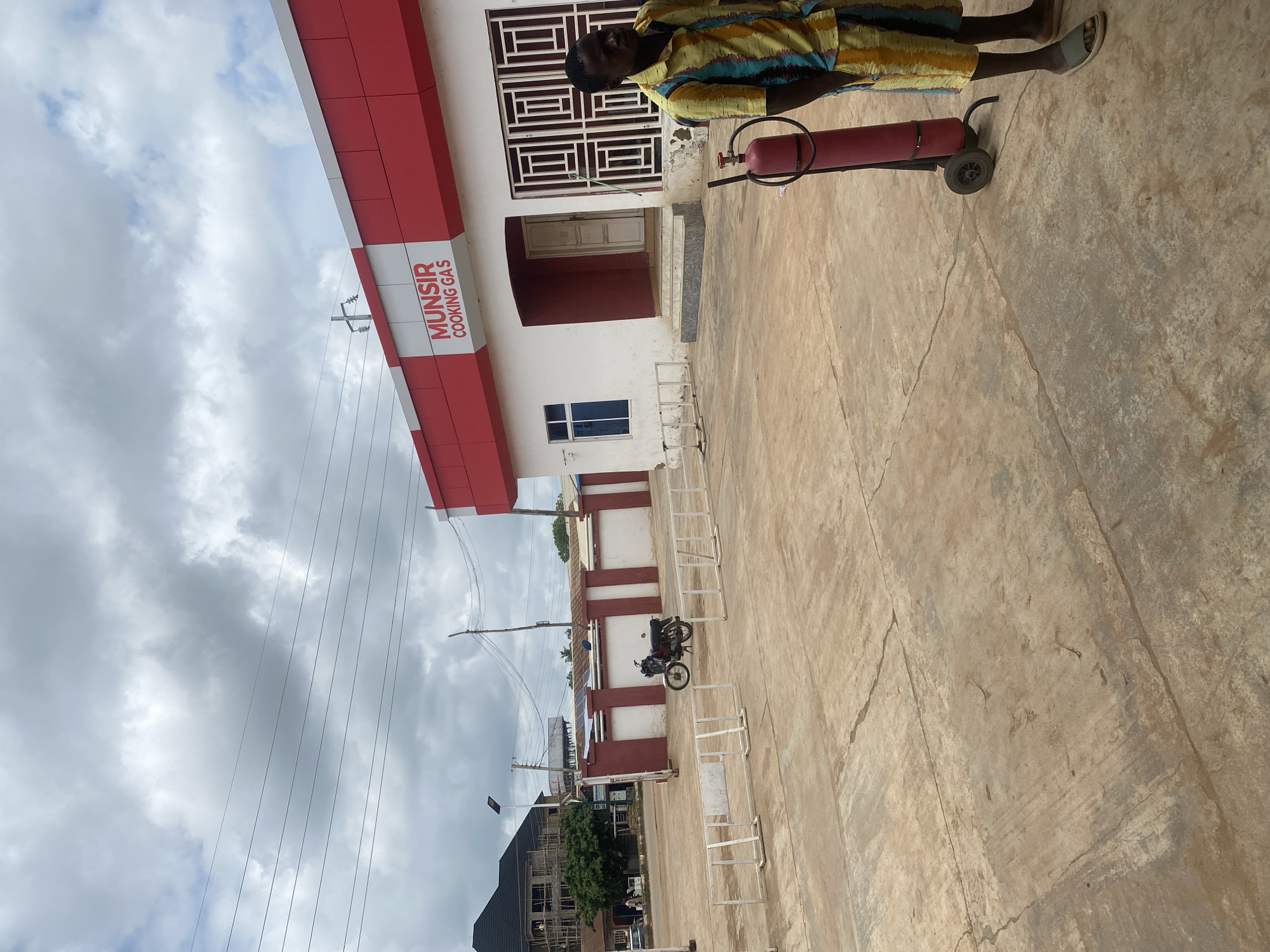 The image shows the exterior of a building labeled "MUNSIR COOKING GAS" in Nassarawa, Nigeria. The building is white with a red trim. In the foreground, a person wearing a colorful striped shirt and shorts stands next to a red gas cylinder on wheels. A motorcycle is parked in the middle ground. The area is paved with concrete, and there are white metal barriers. In the background, a building under construction is visible, along with trees and power lines. The sky is cloudy.