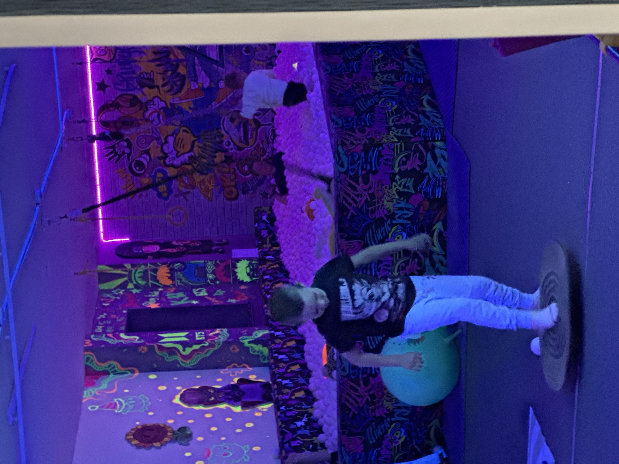 In an indoor recreational space illuminated by purple and blue lights, a young boy stands on a wooden balance board. He is wearing a black t-shirt with a graphic print and light-colored pants. To his right, a large, light green exercise ball is visible. In the background, a ball pit filled with white balls is partially obscured by a colorful mural and a dark fabric barrier. Other children can be seen within the ball pit. The walls of the space are decorated with vibrant, neon-colored graffiti art.