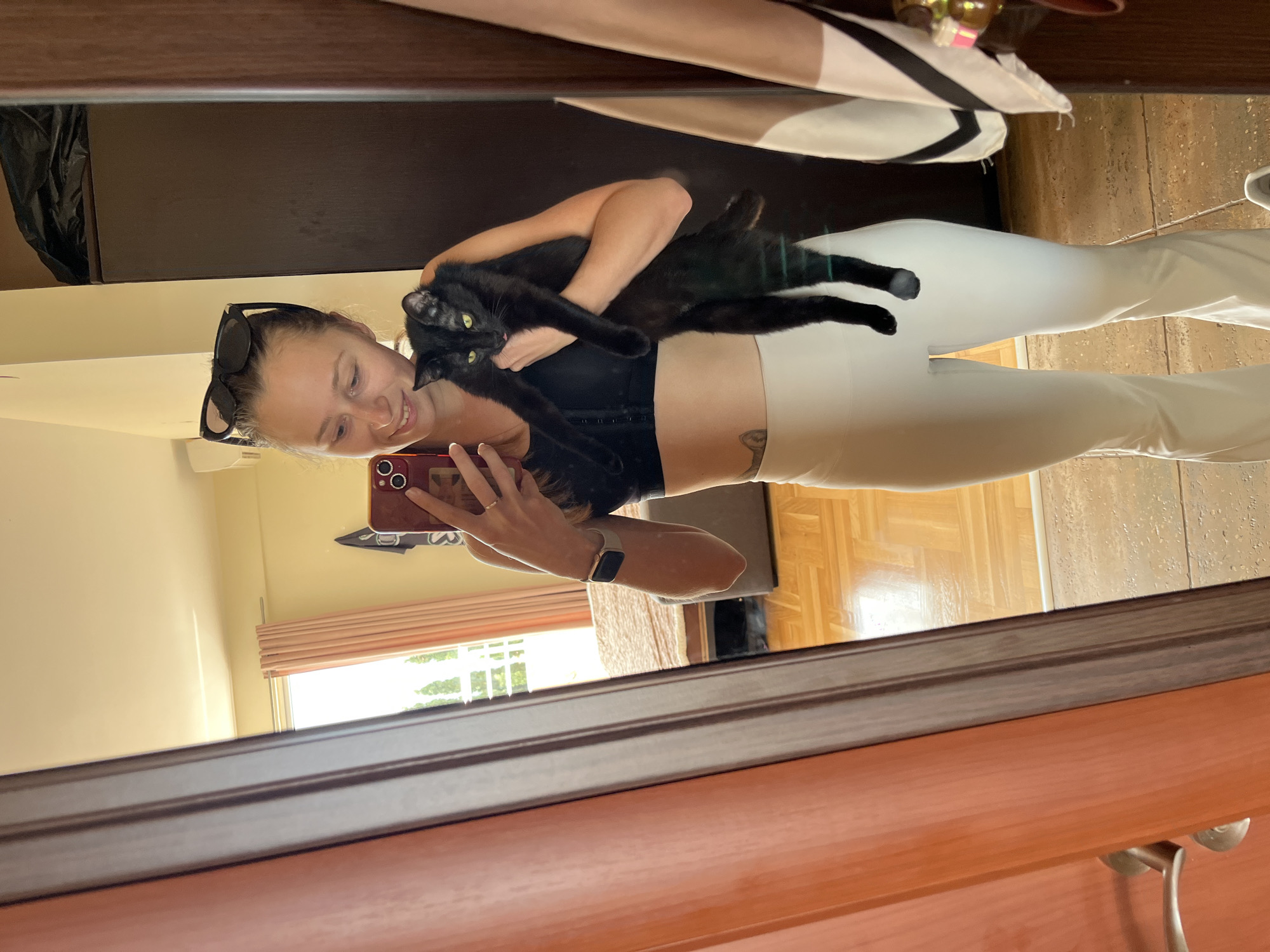 A woman is taking a mirror selfie while holding a black cat. The woman is wearing a black top and white pants, and has sunglasses perched on her head. She is holding a red phone. The cat is sprawled across her arms. The reflection shows a room interior with a window, curtains, and a wooden floor. The mirror frame is brown wood. The location appears to be an interior space, possibly in Bečići, Montenegro, based on the provided context.