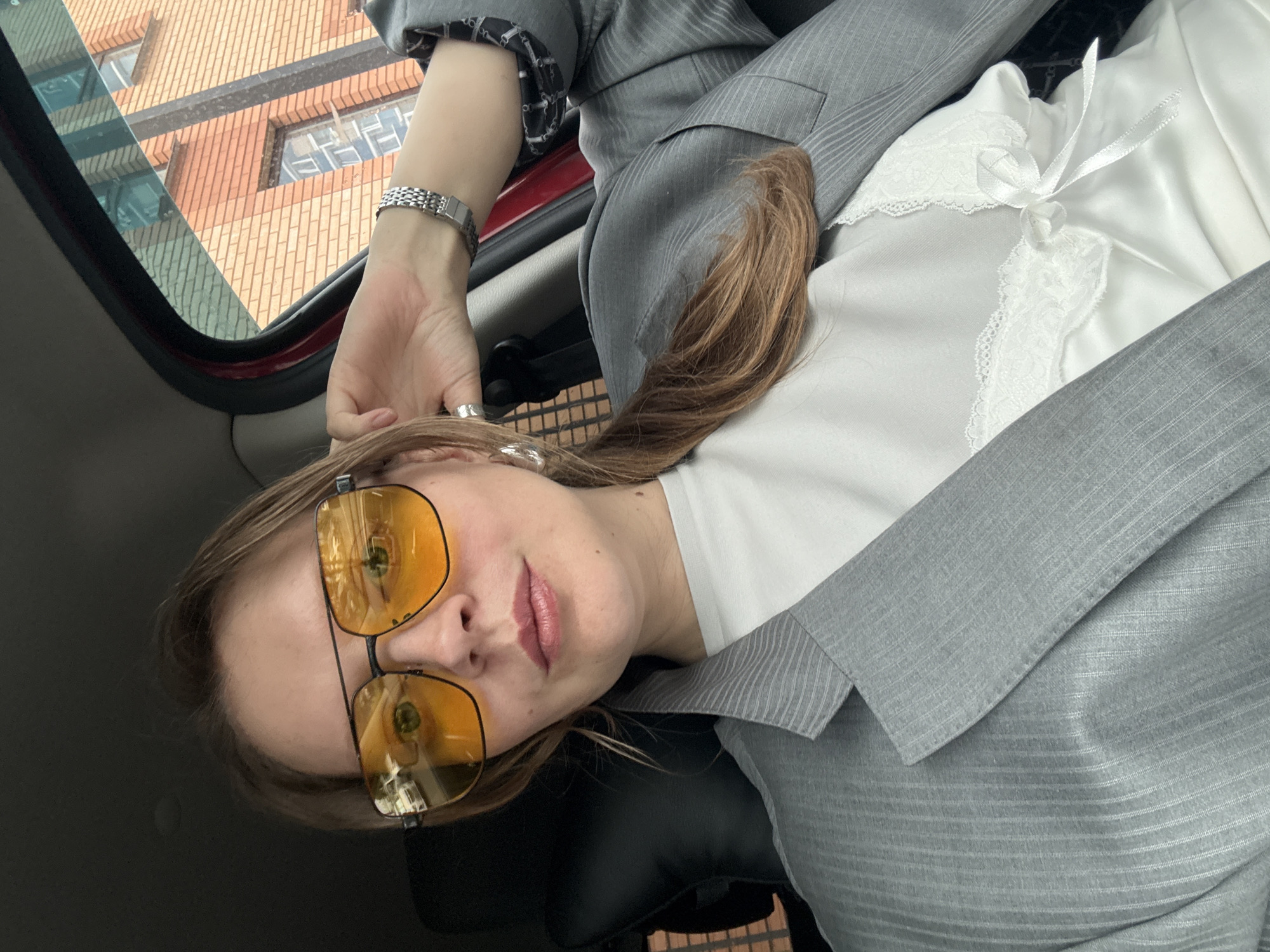 A medium shot shows a woman wearing oversized yellow sunglasses and a grey pinstripe blazer over a white camisole with lace trim, as she sits in the driver's seat of a car. Her right hand is resting on her head, and her hair is pulled back into a ponytail. The interior of the car is visible, with black leather seats and a red frame around the window. Outside the window, a red brick building with windows is visible, suggesting an urban environment. The lighting appears to be natural daylight. This scene is likely taking place in Izhevsk, Russia, given the provided location context.