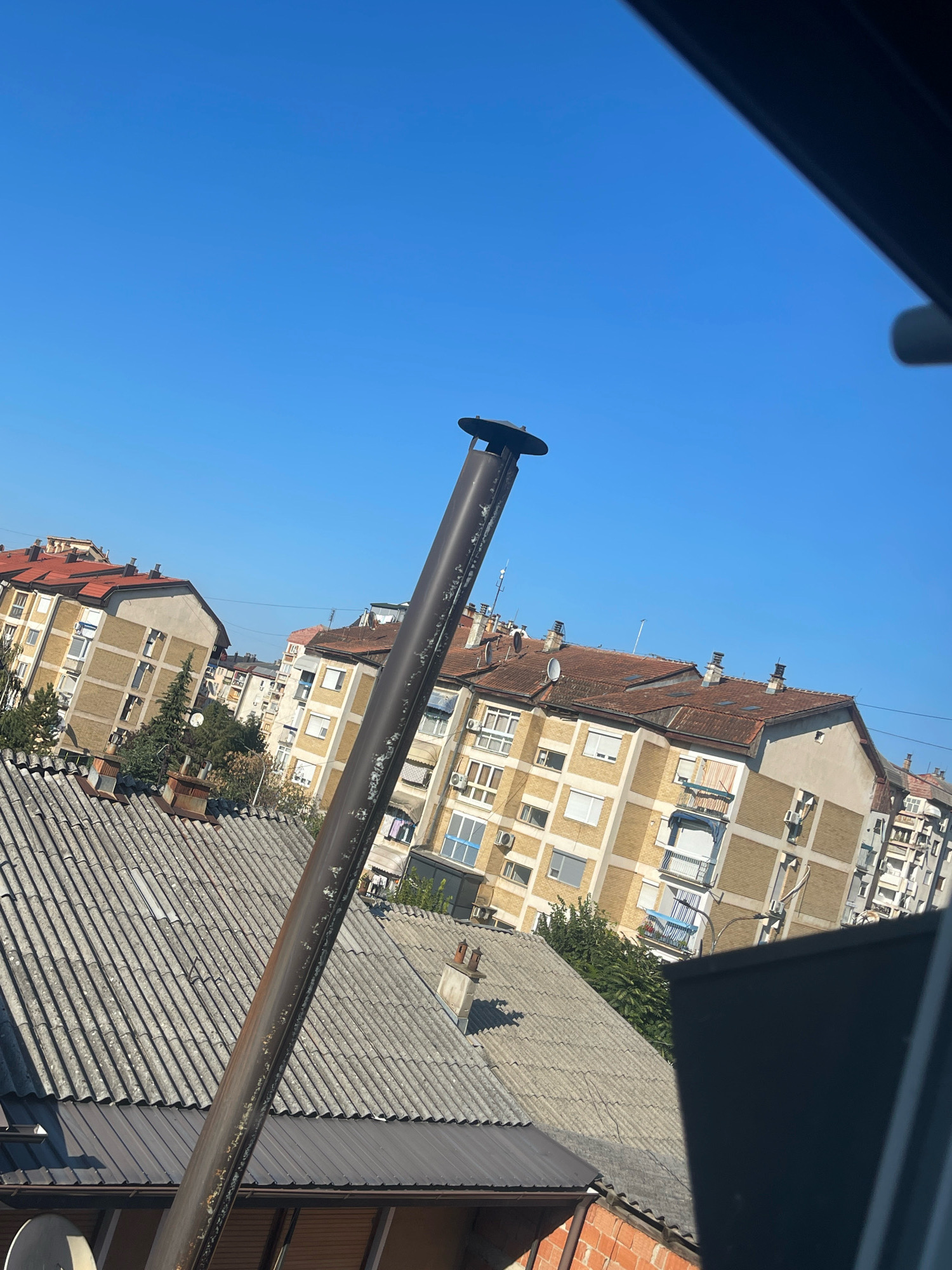 The image captures a view from a window, looking out onto a cityscape under a clear blue sky. In the foreground, a dark, thick metal pipe, likely a chimney or vent, angles diagonally across the frame. It appears weathered and shows signs of rust and wear.
Behind the pipe, several residential buildings are visible. The closest are characterized by grey, corrugated roofs. Further back, taller apartment buildings with light yellow or beige facades and red tiled roofs dominate the scene. These buildings have multiple windows, some with white shutters and balconies. Air conditioning units are visible on some of the building exteriors. Trees with green foliage are interspersed between the buildings, suggesting a residential area with some greenery.
The perspective suggests the photograph was taken from an elevated position, possibly a higher floor of a building. The angle of the buildings and the pipe suggests the camera may be tilted. The overall impression is of an urban environment on a sunny day. The specific location in Prizren, Kosovo, is not directly indicated by any visual cues in the image itself.