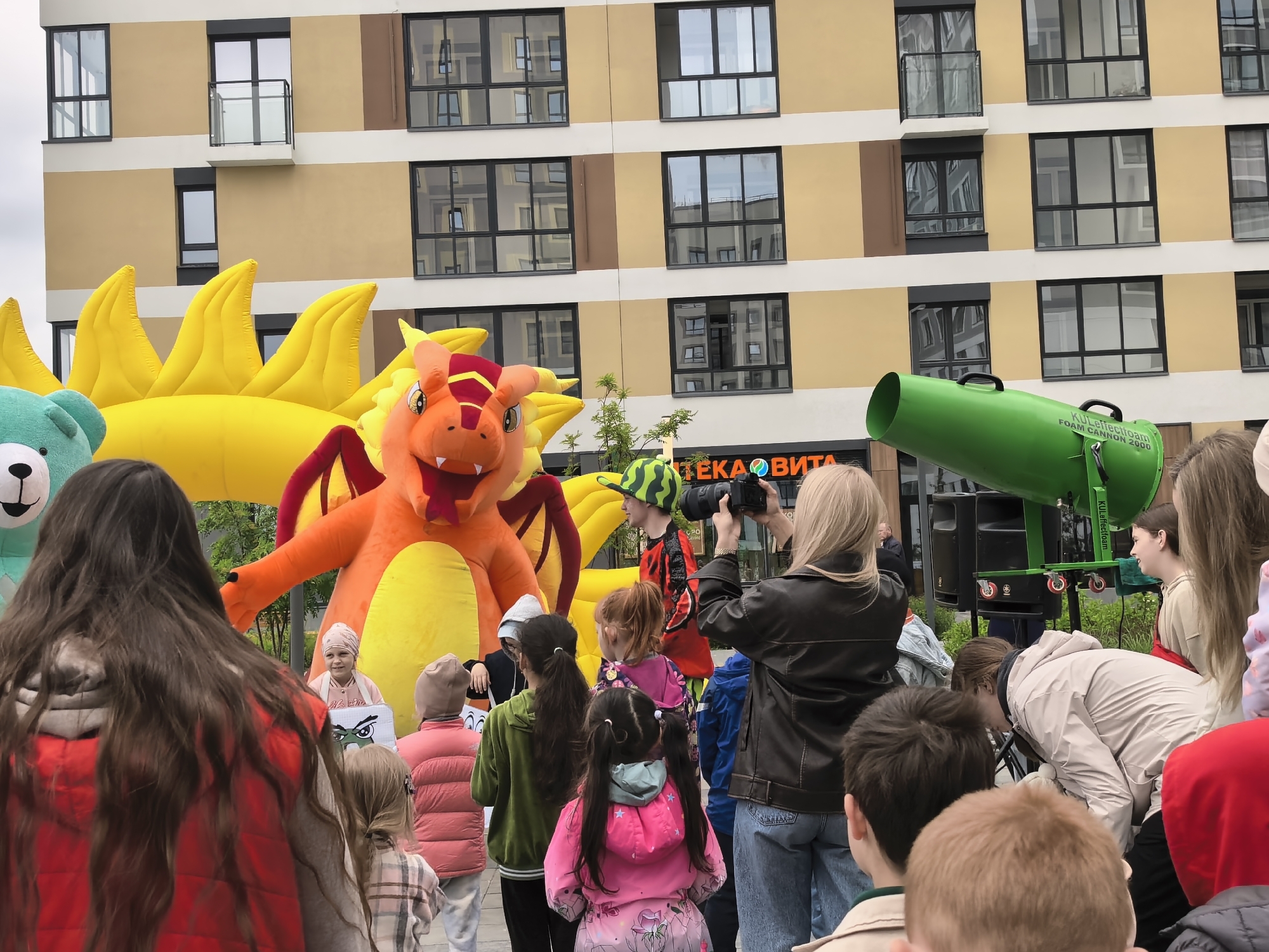 The image shows a gathering of people, predominantly children, in what appears to be an urban event. In the background is a multi-story building with a beige facade and dark-framed windows. A large inflatable figure resembling a dragon is centrally positioned, along with another inflatable figure of a blue bear. A person wearing a costume with a watermelon-patterned hat is present near a woman holding a camera, who seems to be documenting the event. To the right of the frame, there's a green object that looks like a foam cannon labeled "KULJetectofoam FOAM CANNON 2000". The crowd of people is varied in age and attire, suggesting a family-oriented event. A storefront with the text "TEKA BITA" is visible behind the woman with the camera. The location is Unknown, Unknown.